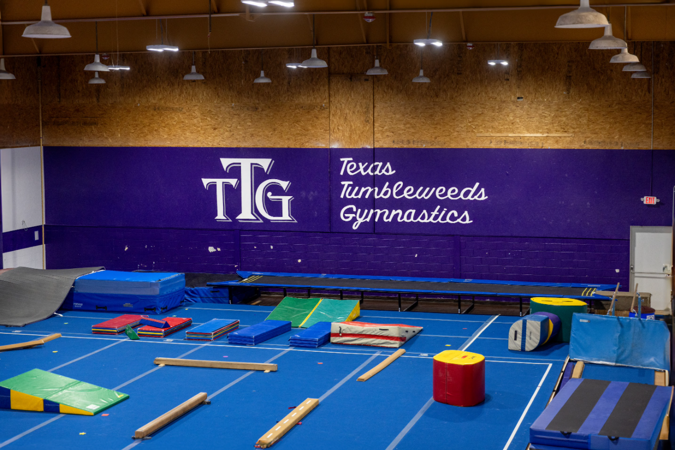 Interior of a Texas Tumbleweeds Gymnastics facility with mats, equipment, and the company logo on a large purple wall.