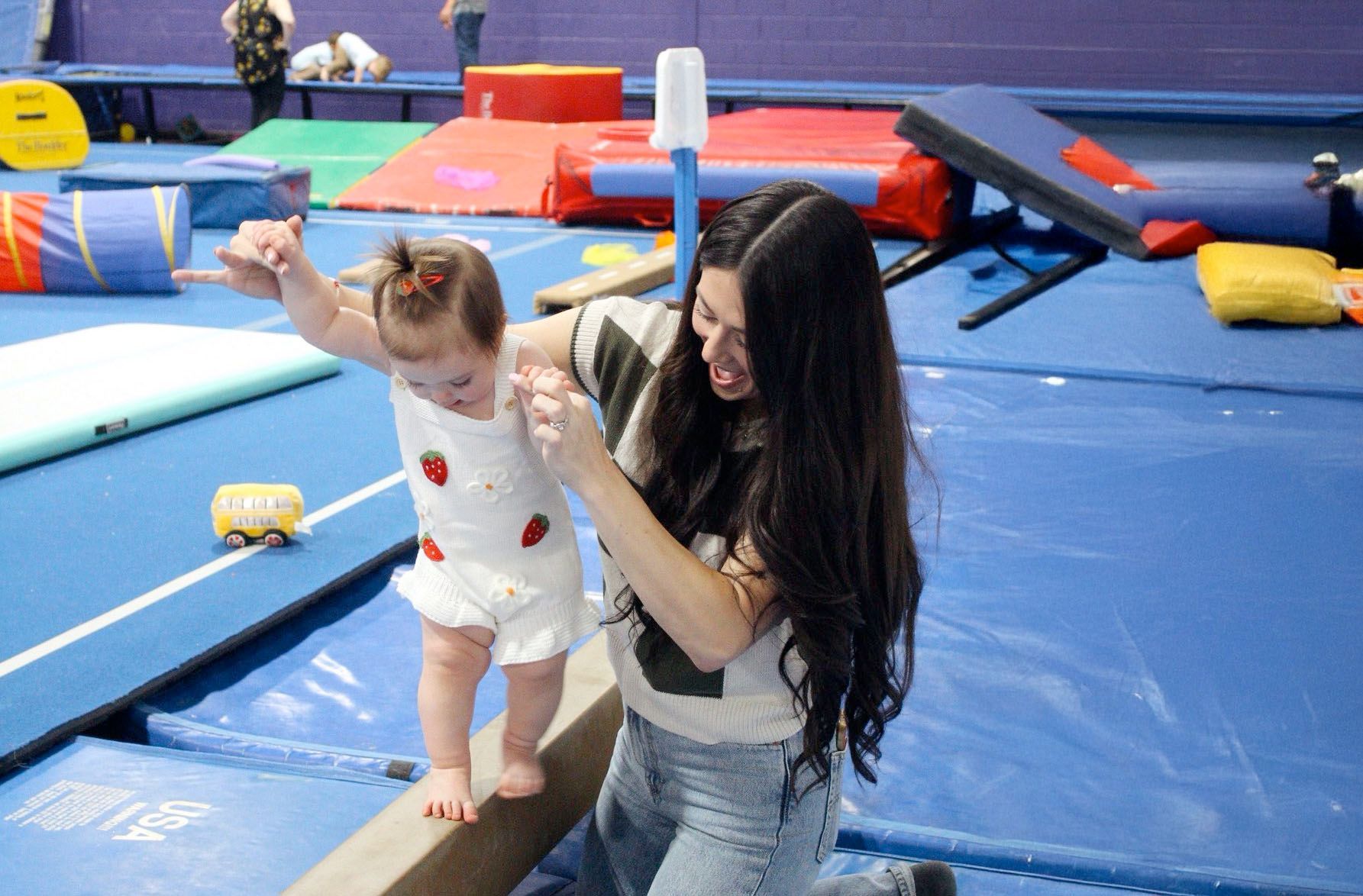 A person helps a young child walk across a balance beam in a brightly colored gymnastics studio.