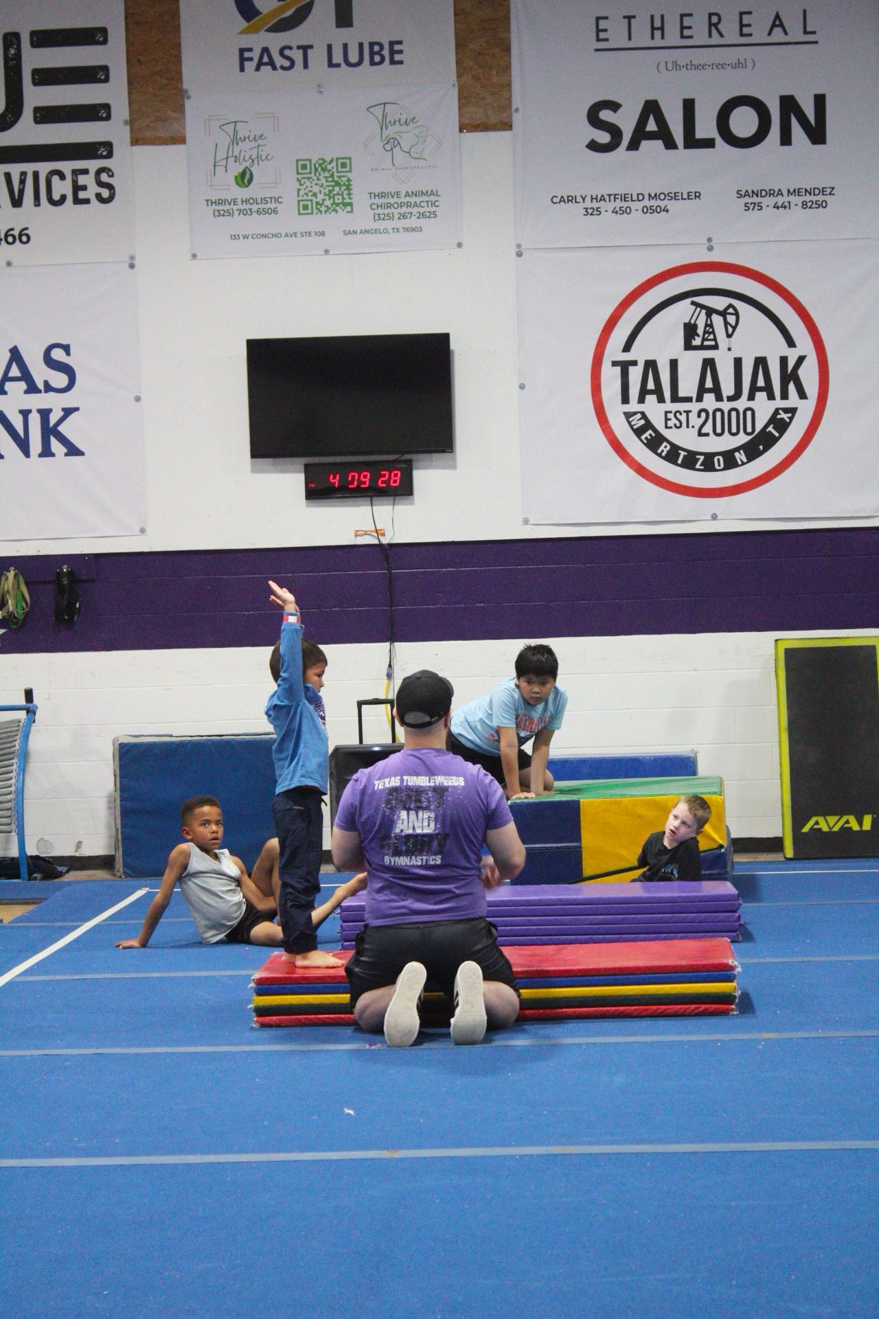 An instructor kneels on a blue mat in a gym, guiding a student with raised arms, while others wait nearby.
