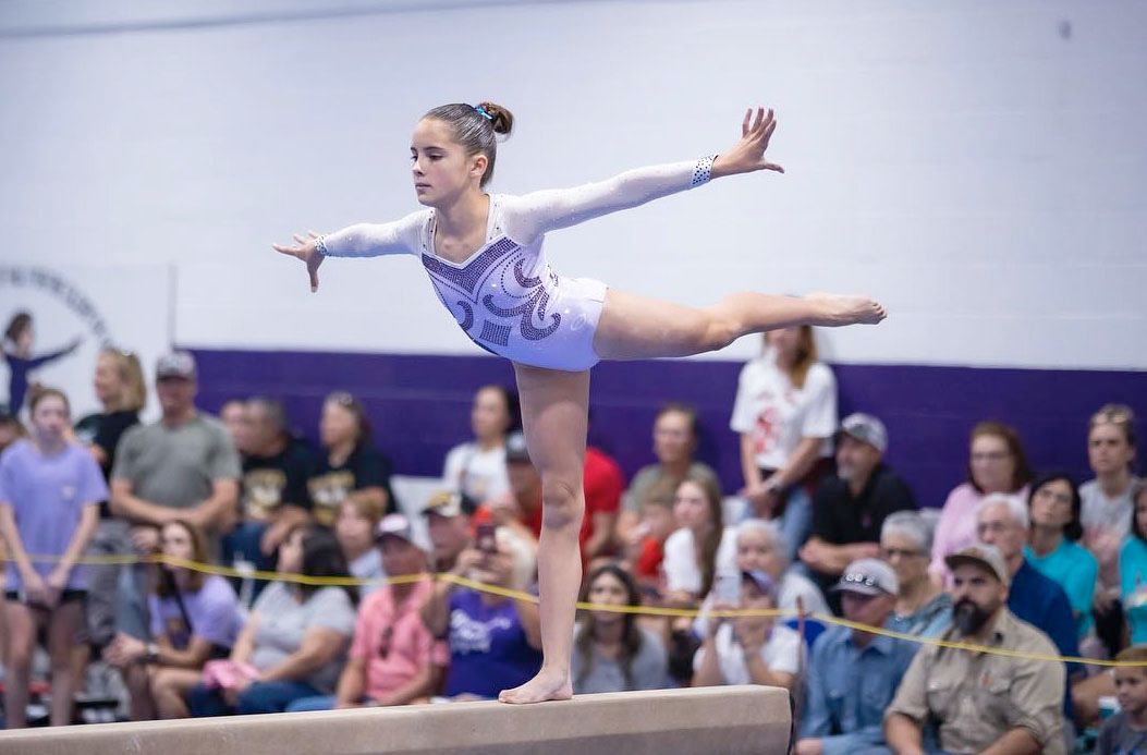 A gymnast in a white and purple leotard performs a balance skill on a beam in front of an audience.