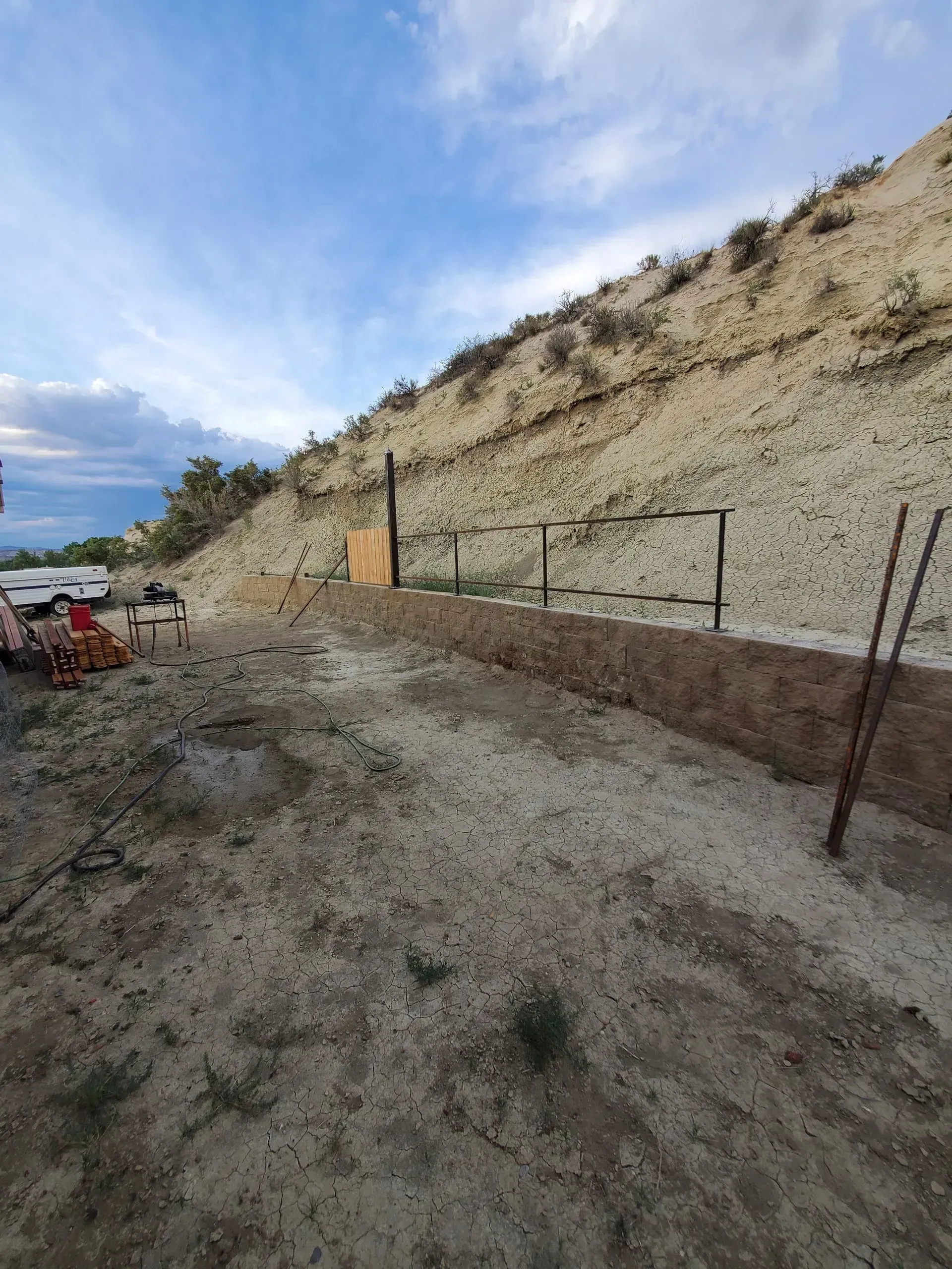 Dirt road beside a steep rocky hillside under a cloudy blue sky, with a fence and a small gate.