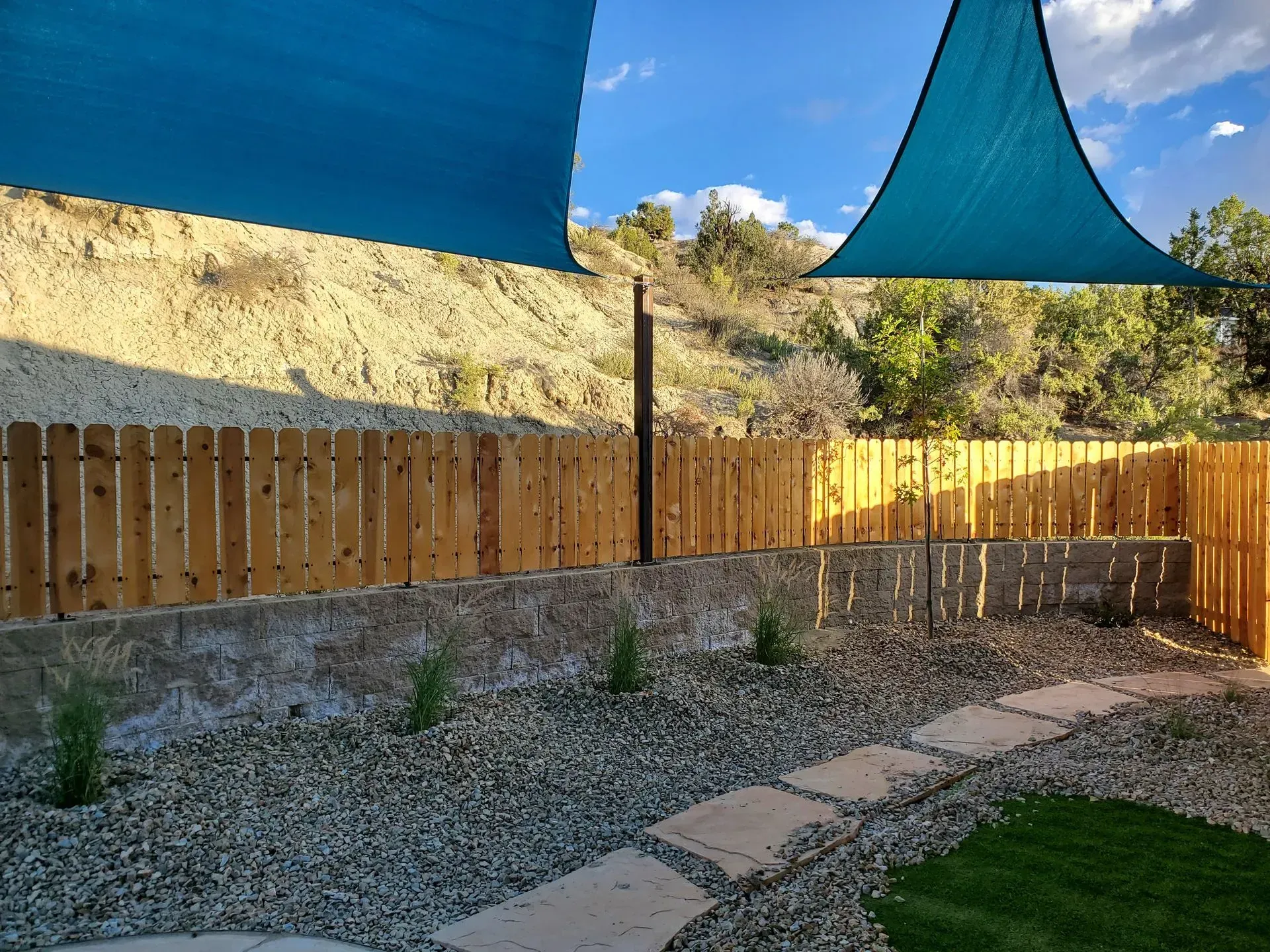 Backyard patio with wooden fence, gravel, stone path, and blue shade sails over a grassy corner