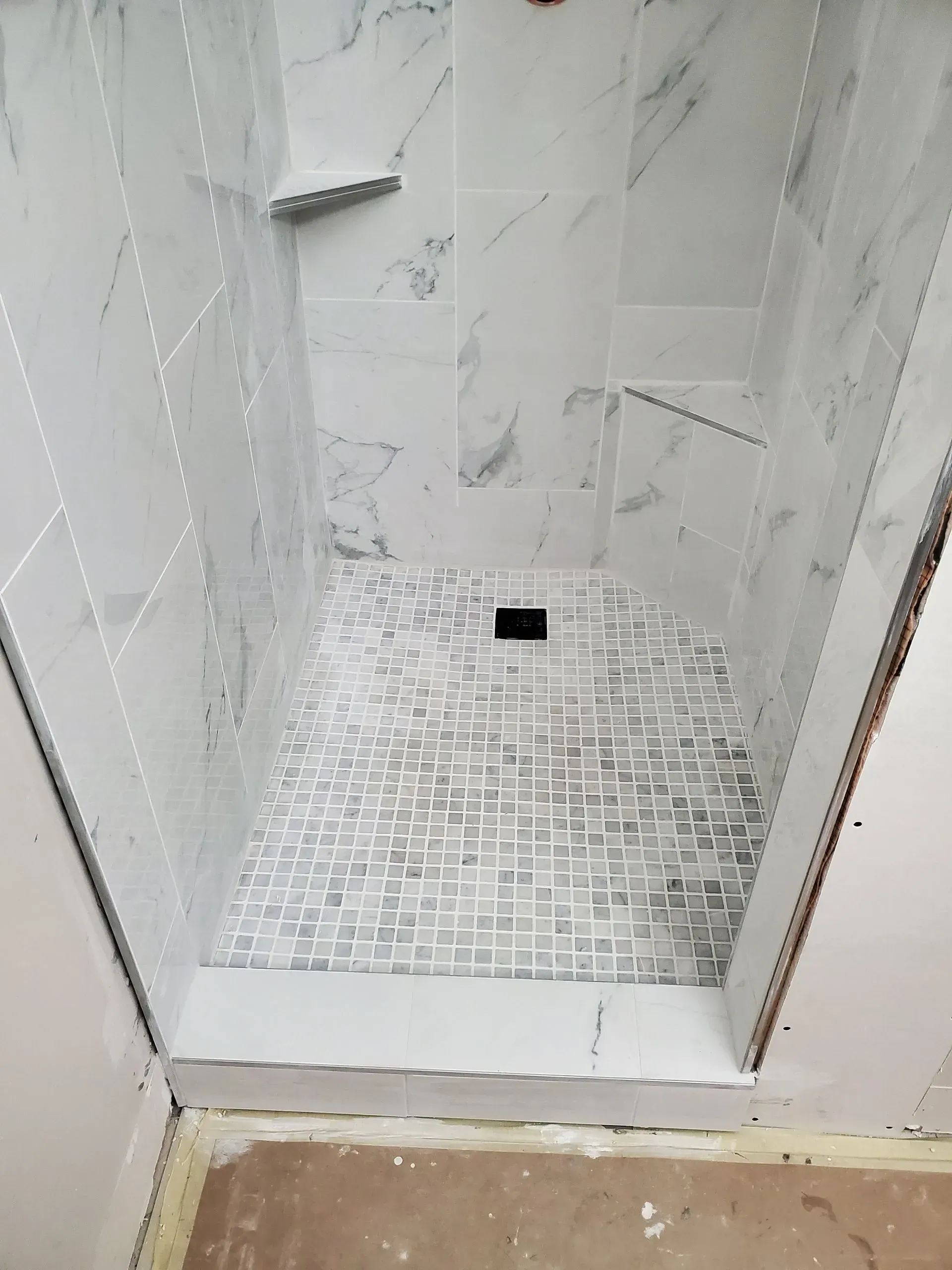 Tiled shower stall with a small shelf and floor drain, viewed from the bathroom entrance.