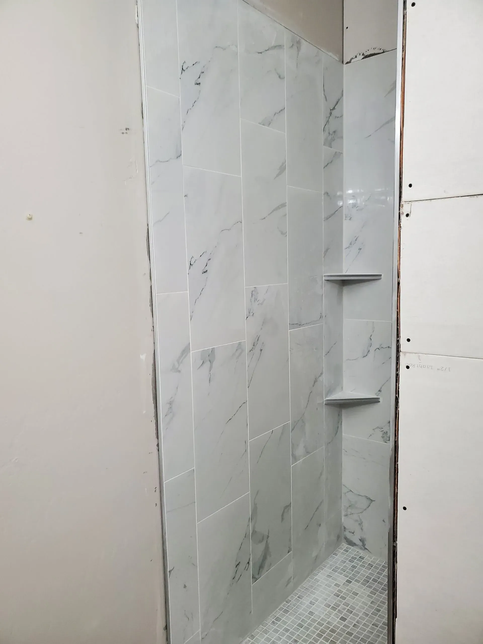 Narrow unfinished bathroom shower stall with gray tile, shelves, and exposed gravel floor