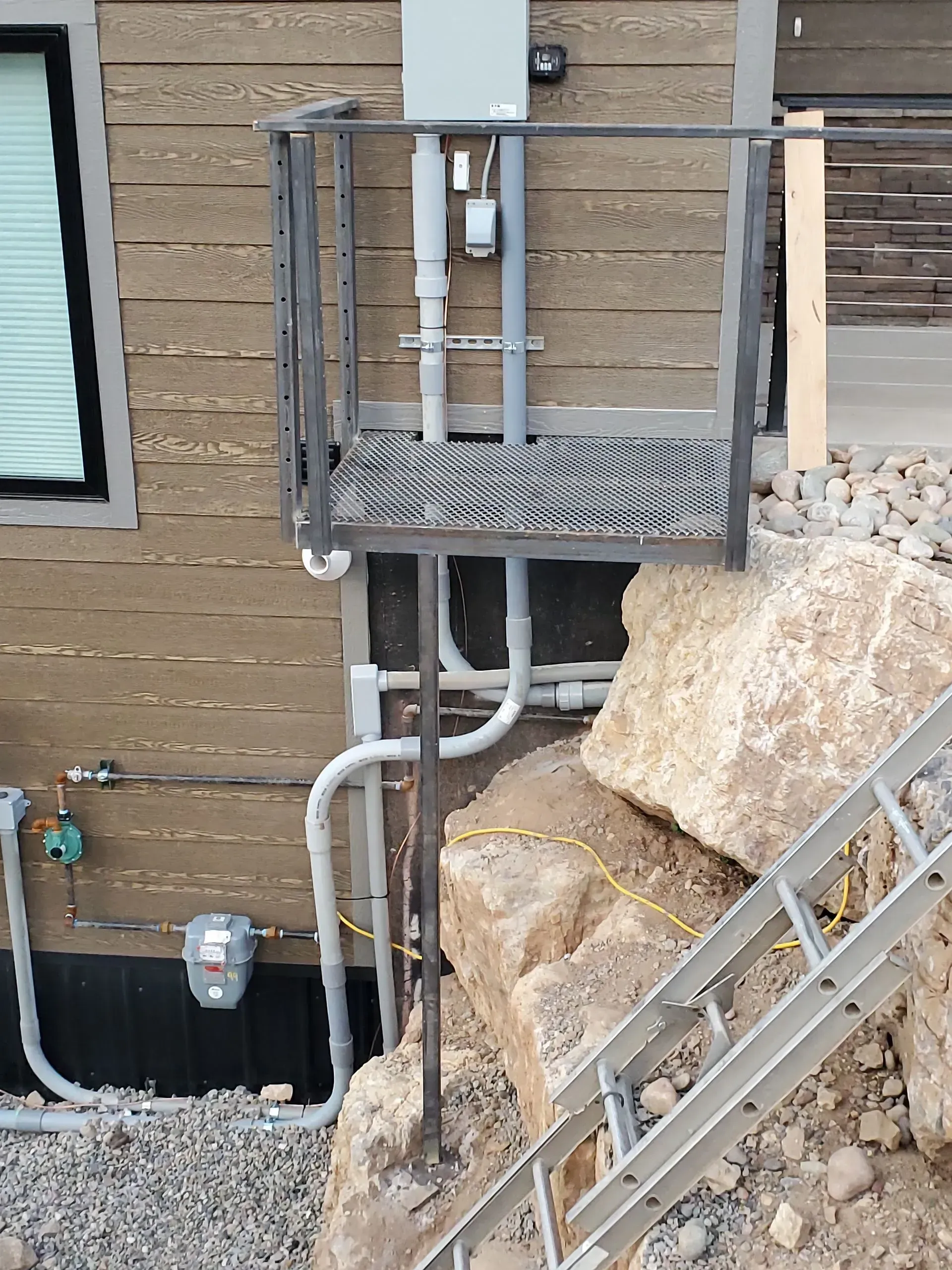 Exterior utility platform and piping beside a brick building, with rubble and a metal ladder in front.