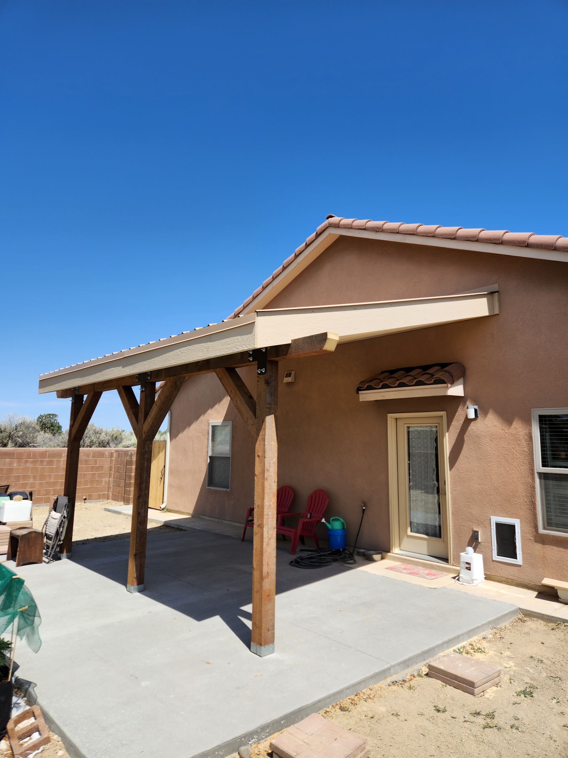 Desert home patio with wooden pergola, concrete slab, and tan stucco wall under a clear blue sky