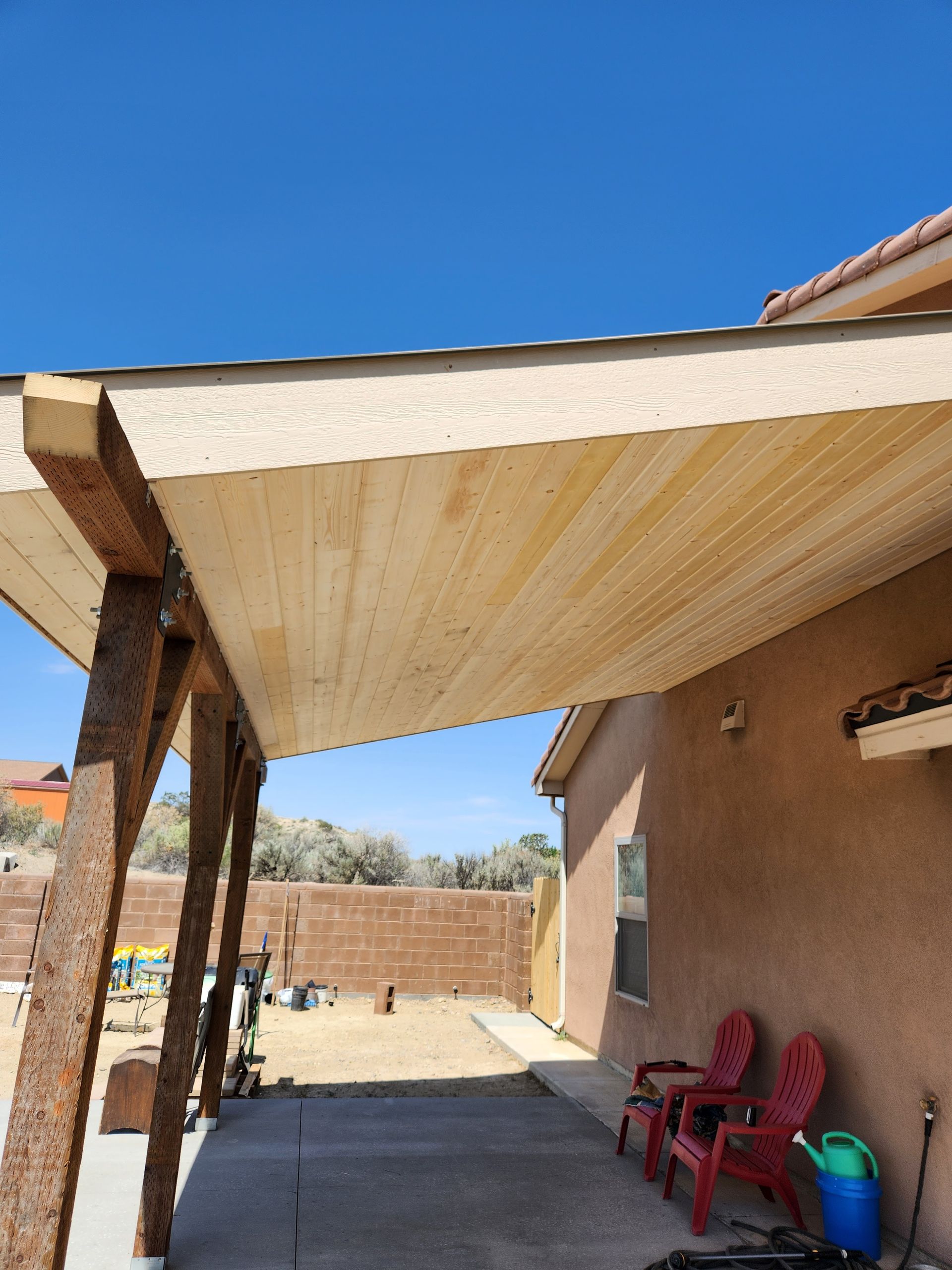 Covered patio with a slanted roof, brick posts, red chairs, and a blue sky over a yard wall