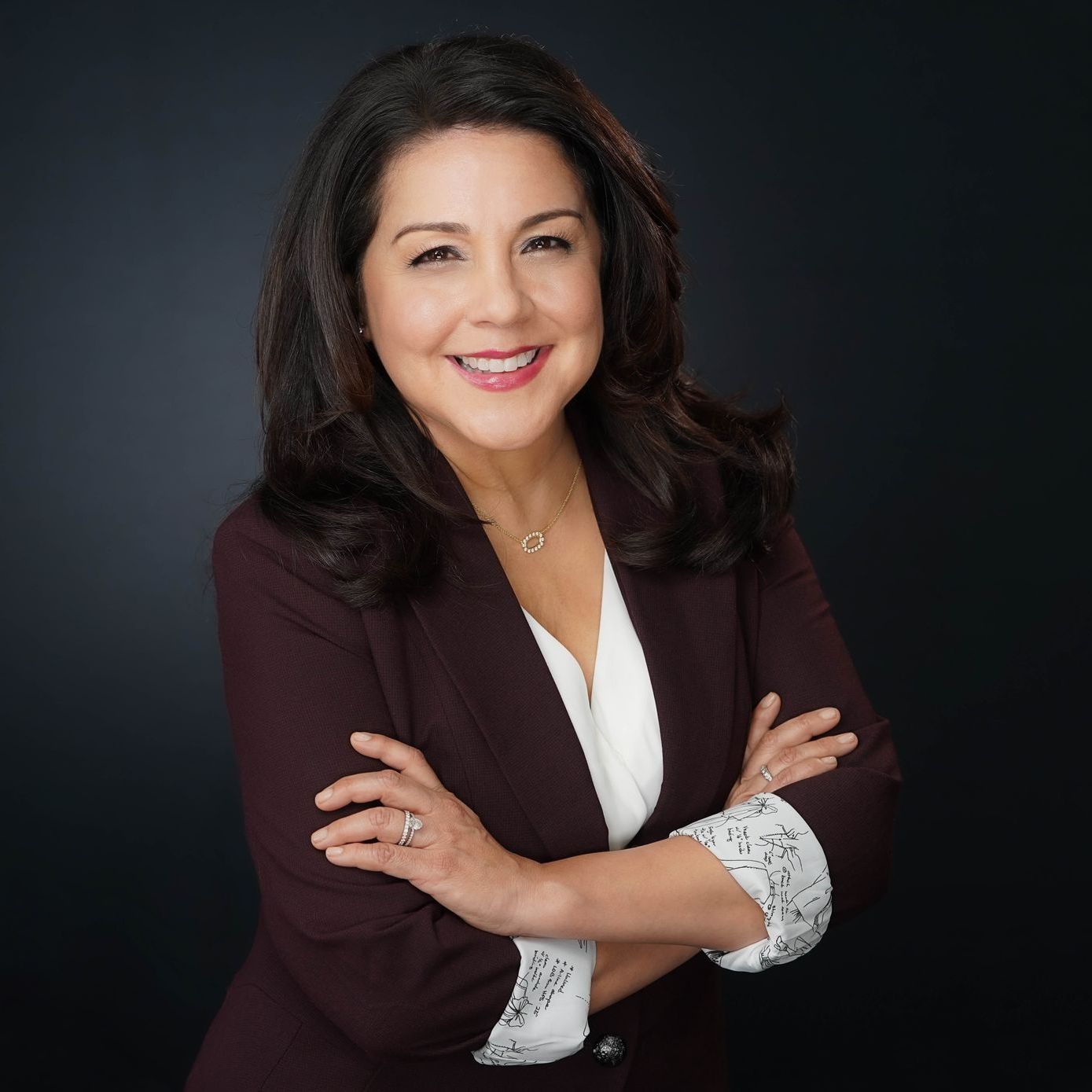 A woman in a burgundy jacket and white shirt is smiling with her arms crossed.