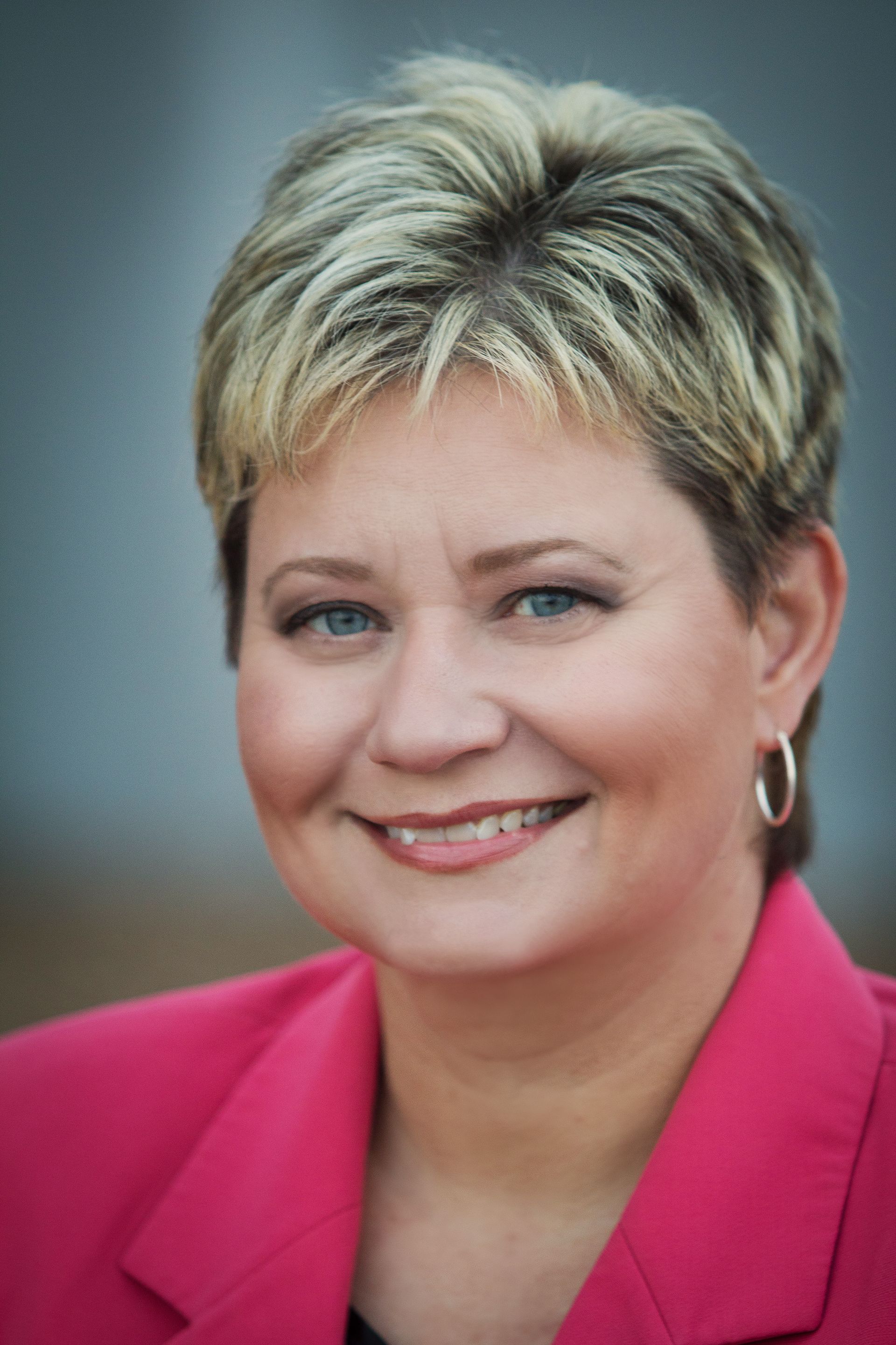 A woman wearing a pink jacket and hoop earrings is smiling for the camera.