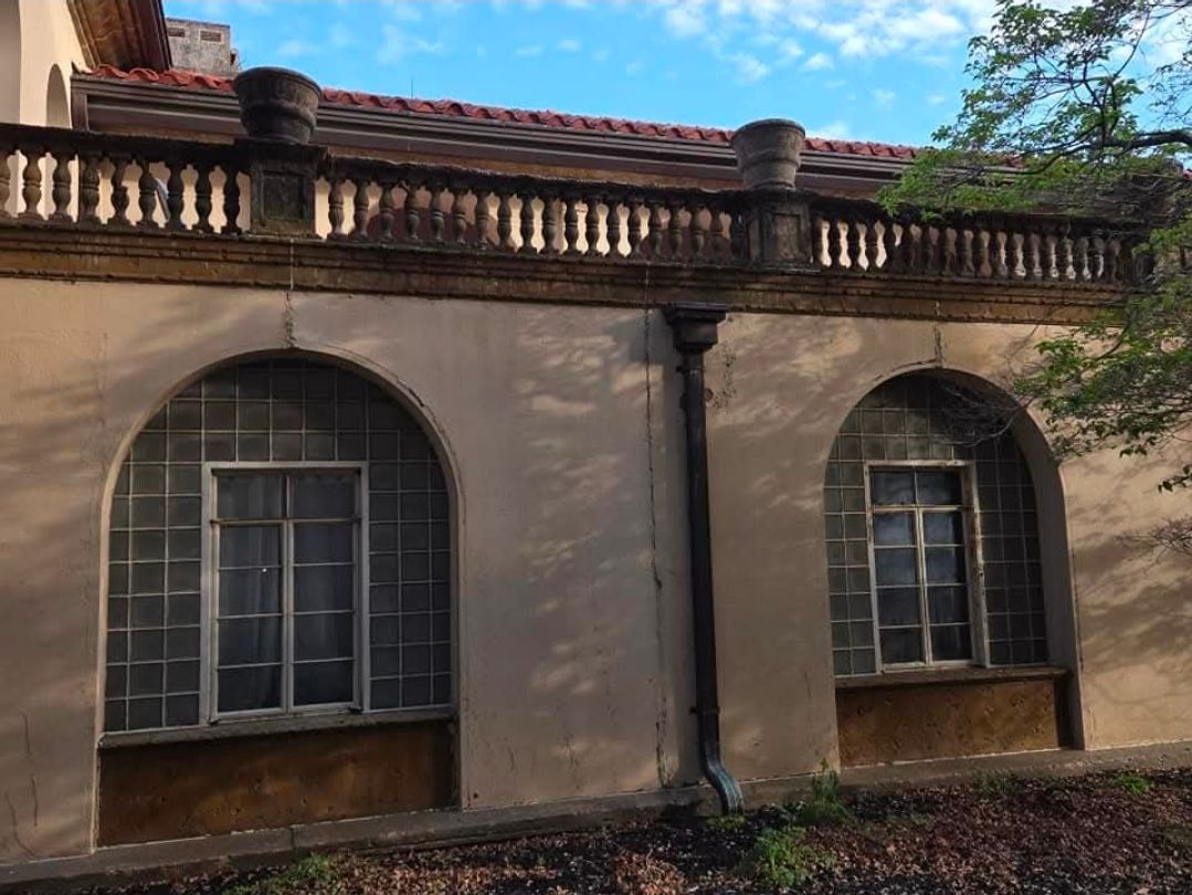 Beige building exterior with two arched, grid-paned windows, a stone balustrade roof terrace, and a brown downspout.
