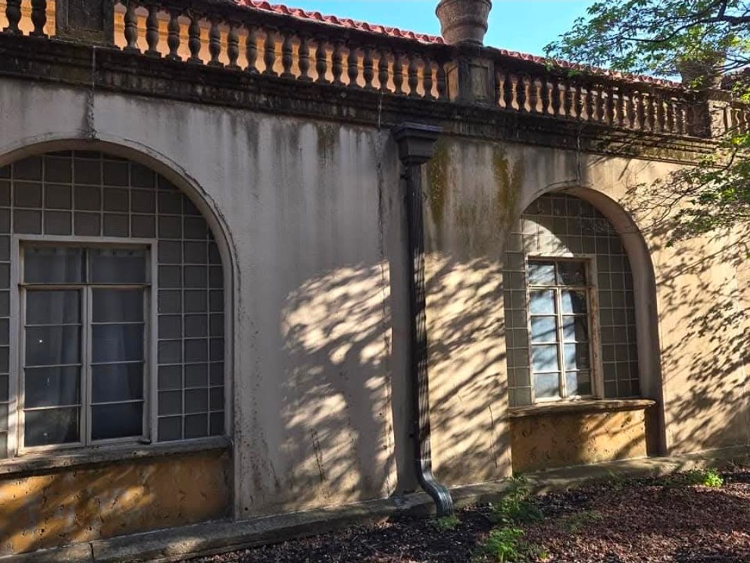 Old stucco building exterior with two arched windows, a metal drainpipe, and a decorative balustrade along the roofline.
