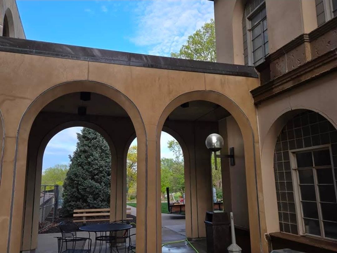 An outdoor patio with tan arched walkways, a round table with chairs, a metal lamp post, and trees in the background.