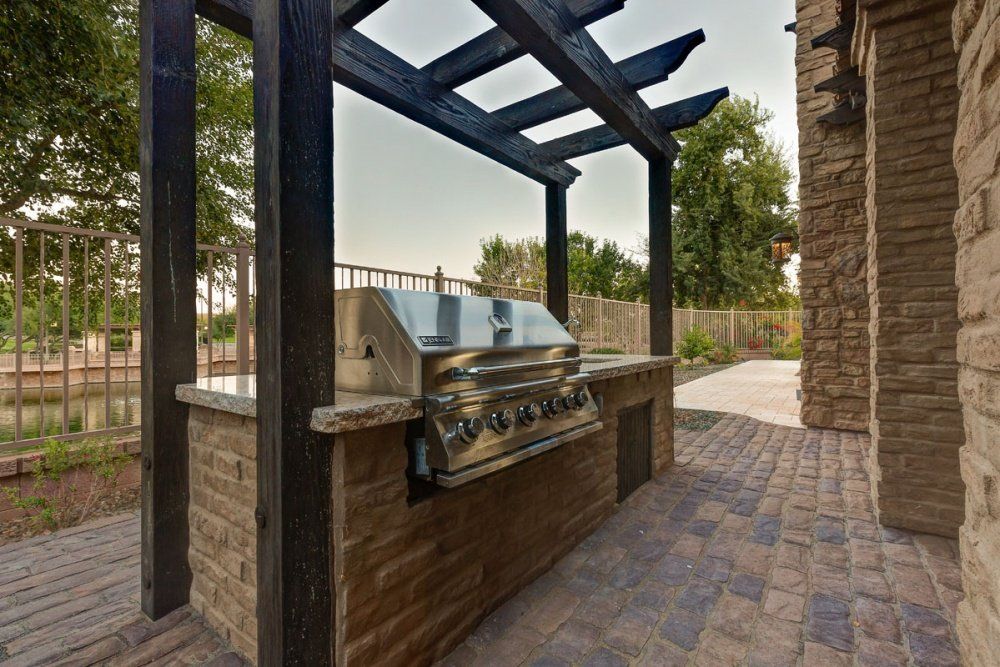 A grill is sitting under a pergola on a brick patio.