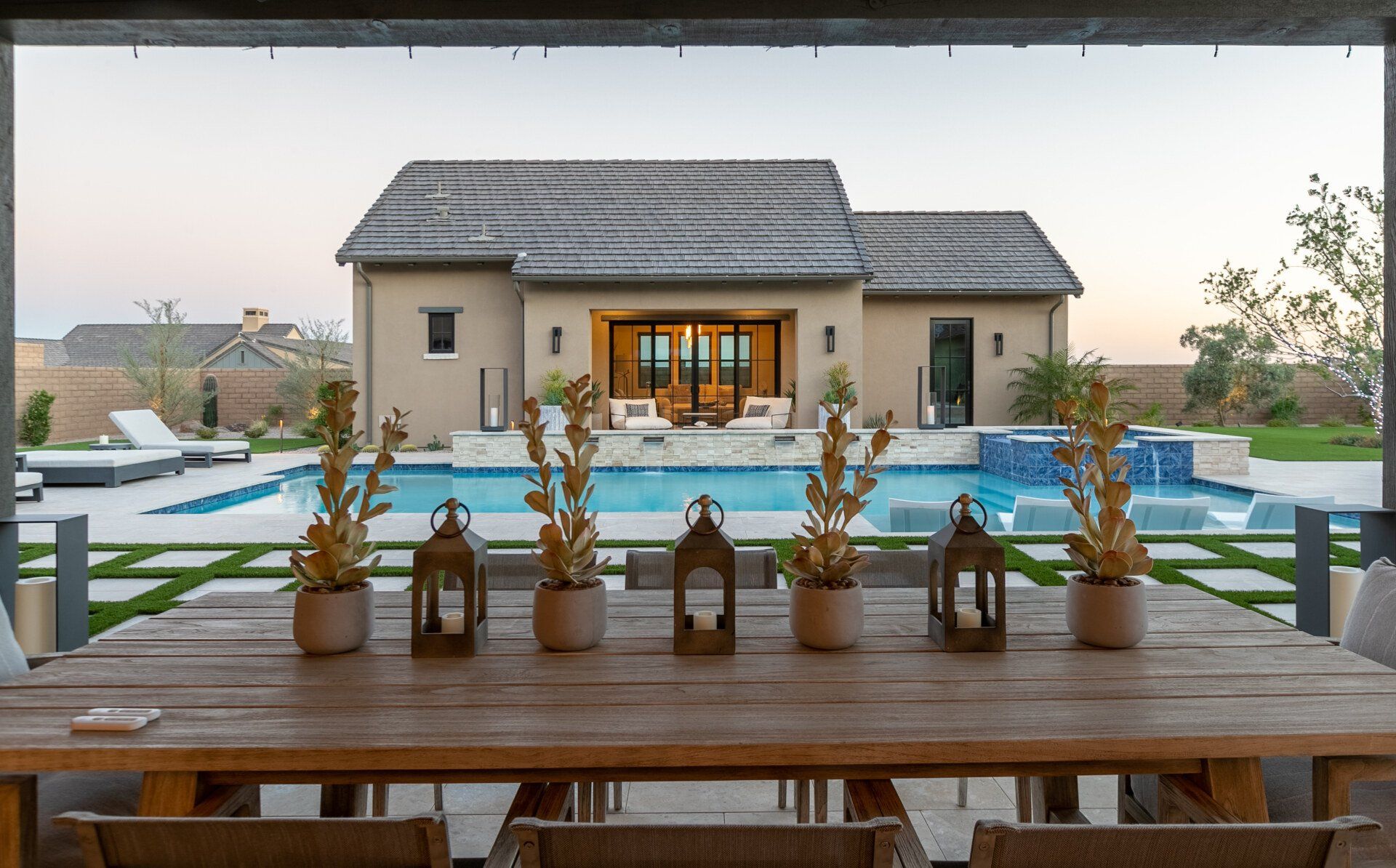 A dining table with lanterns on it in front of a house with a pool.