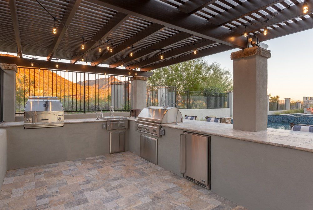 A large outdoor kitchen with a grill , sink , and refrigerator under a pergola.