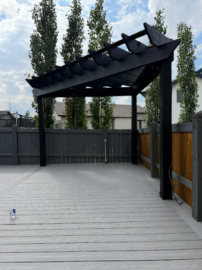 Black pergola on a gray wooden deck, with a gray fence and trees in the background.