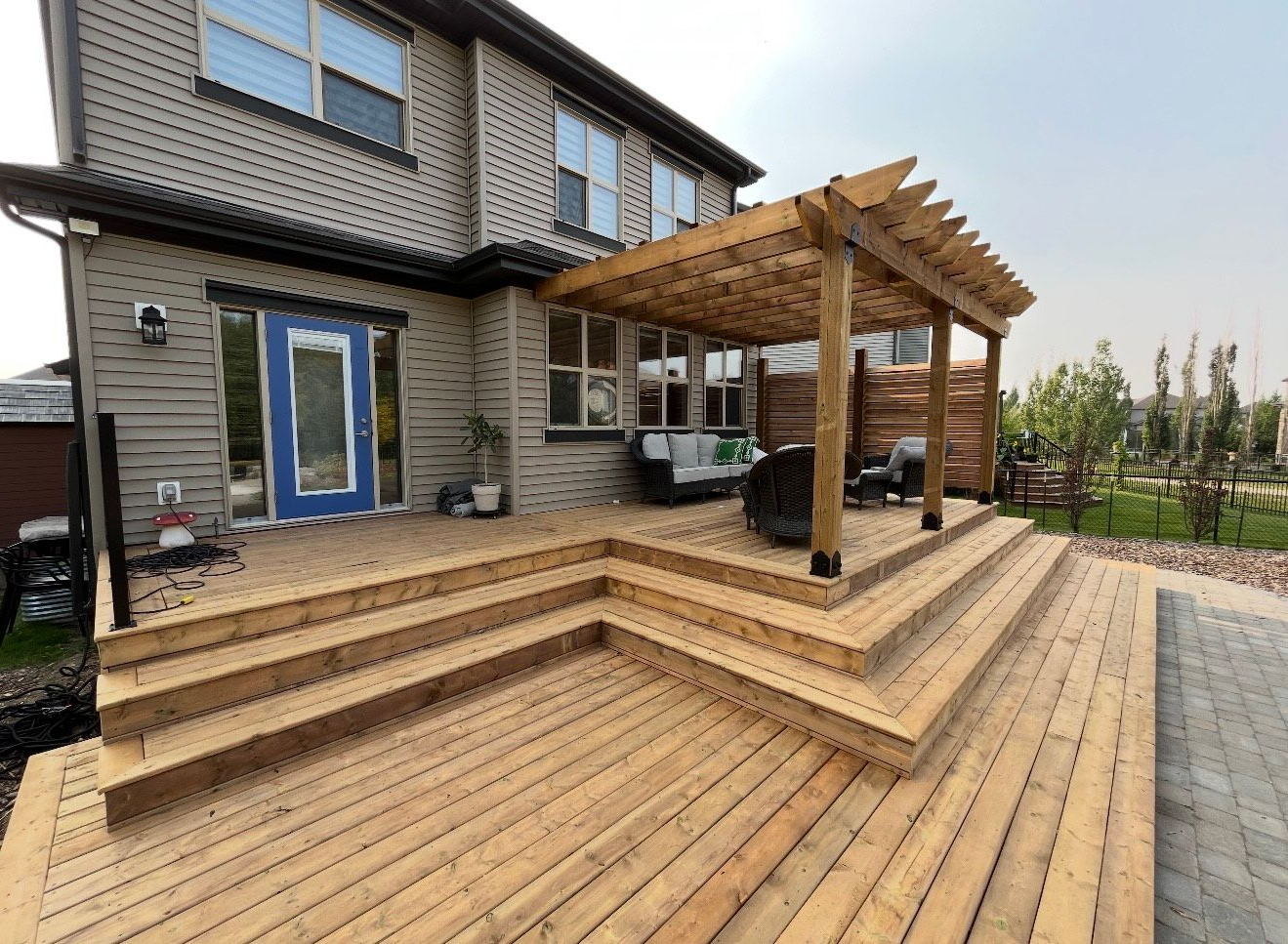 Wooden deck with steps leading to a pergola attached to a two-story brick house with a blue door.