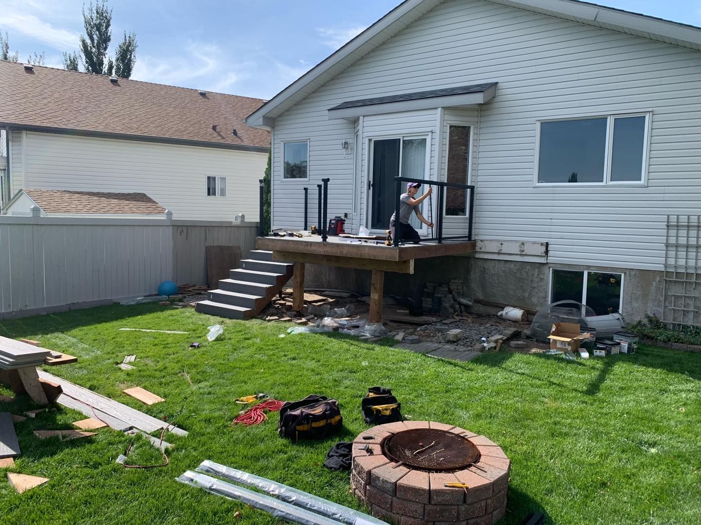 Backyard deck under construction; person on deck, tools, fire pit, white house, green grass, light sky.