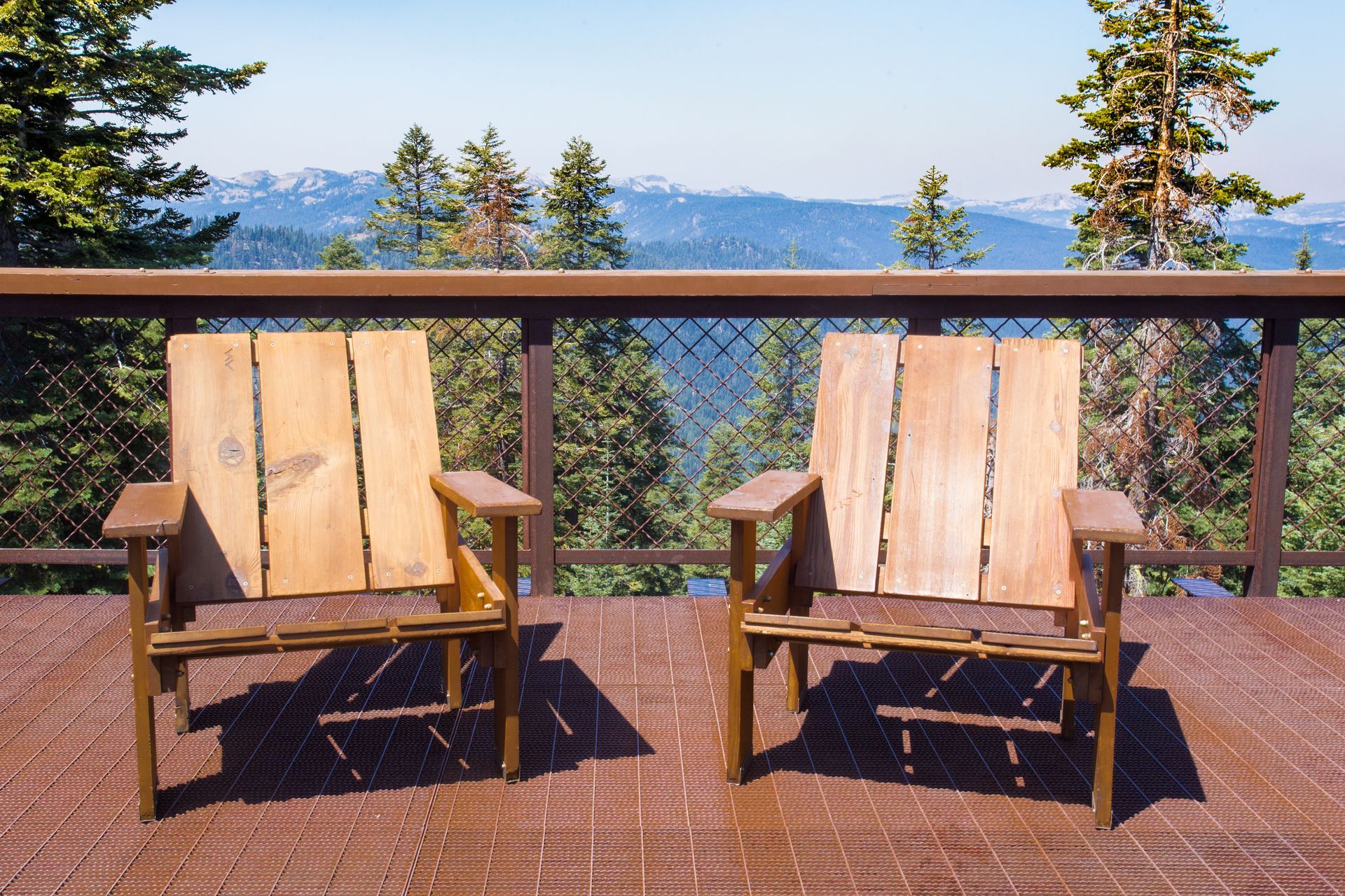 Two wooden chairs on a deck overlook a forest and mountain range on a sunny day.