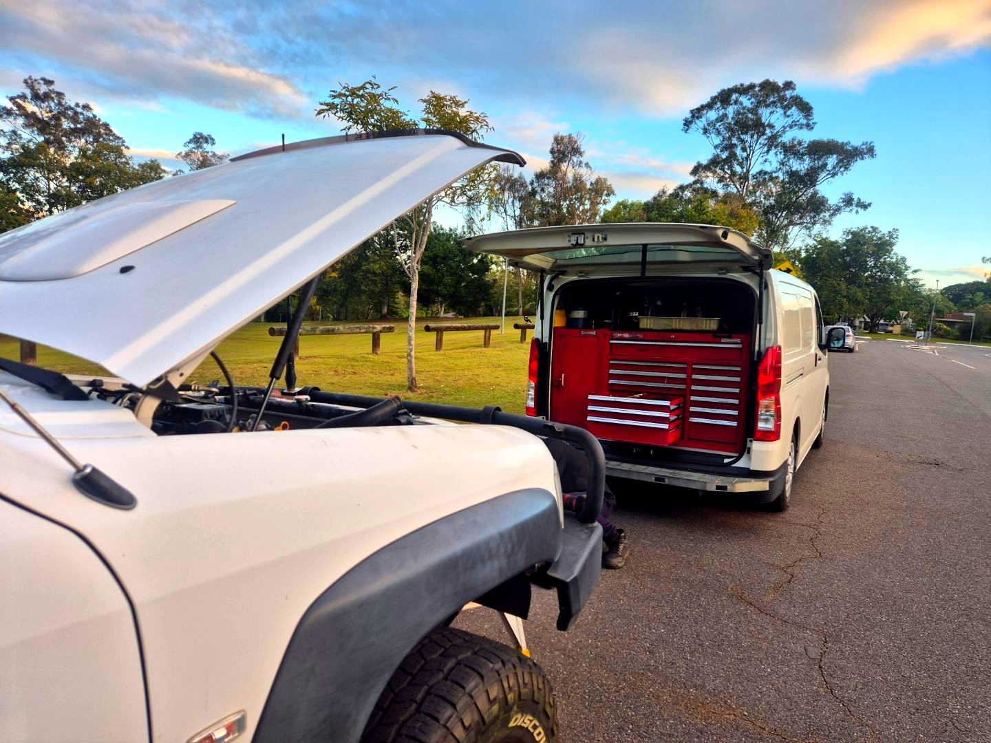 White SUV with hood up, next to white van with open back holding a red toolbox, parked on a road.