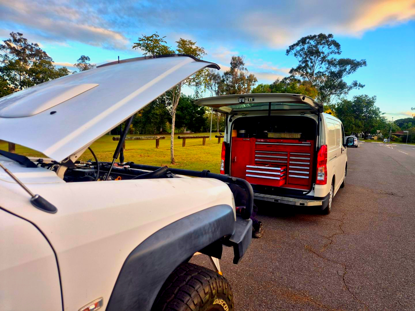 White truck with hood up next to a white service van with open doors, tools visible, parked on a road.