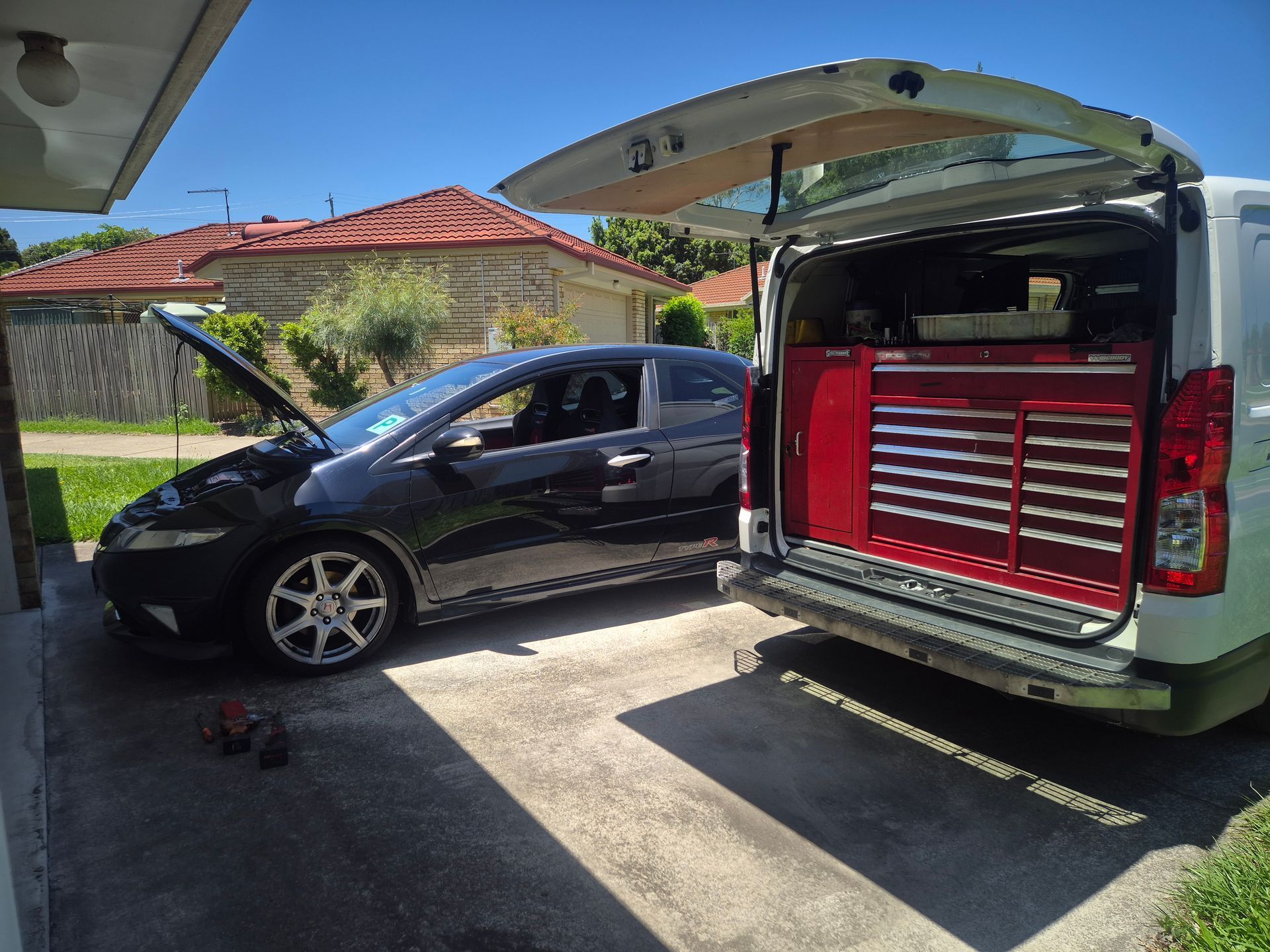 Black car with open hood next to a white van with open back, revealing a red tool chest. Outdoors.