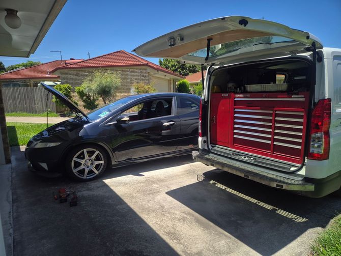Black car with open hood next to a white van with open back, revealing a red toolbox. Outdoors, sunny.