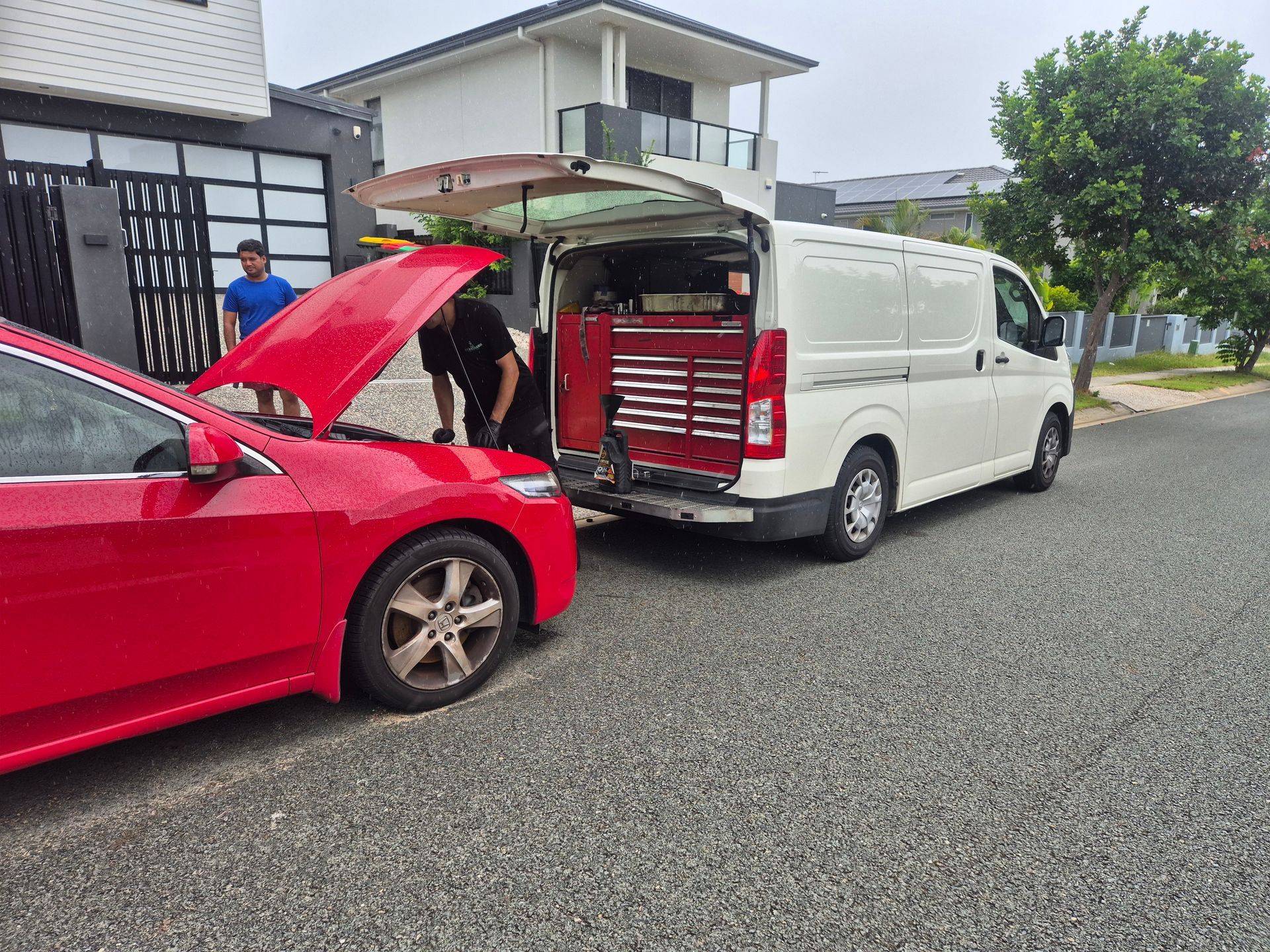 Red car and white van with an open rear hatch, showing tools in the back on a residential street.