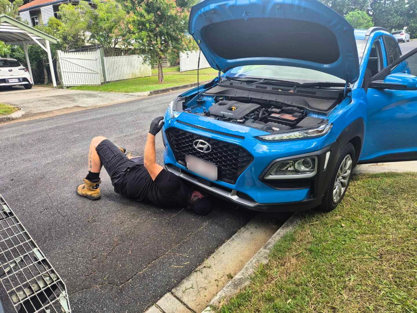 A mechanic lies under a blue car with the hood open, working on the engine. They're on a street next to a curb.