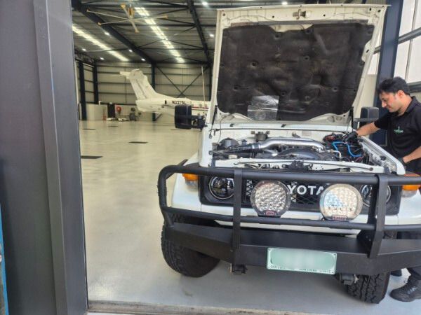 White Toyota Land Cruiser with open hood being worked on, in a hangar with a plane in the background.