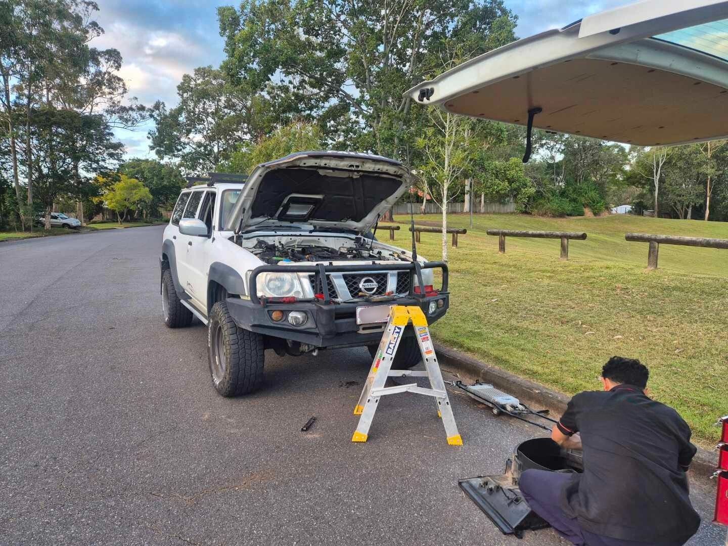 White pickup with hood open on roadside; person kneels nearby working with tools and a step ladder.