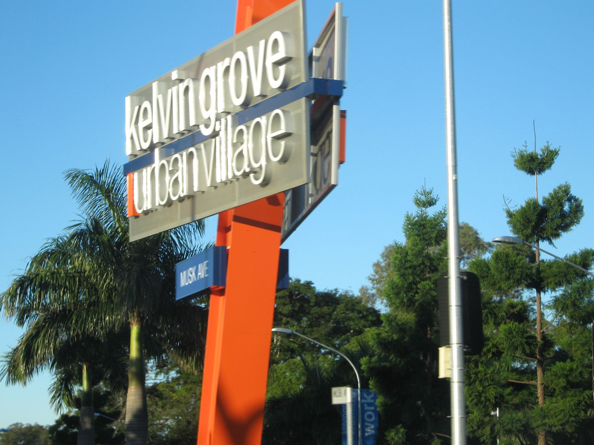 Kelvin Grove Urban Village sign, orange pole, blue accents, silver letters, trees and blue sky background.