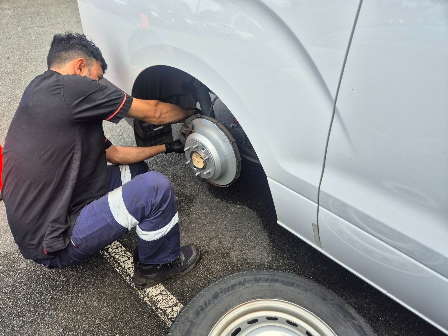 Mechanic changing a tire on a white van outdoors, wearing work clothes.