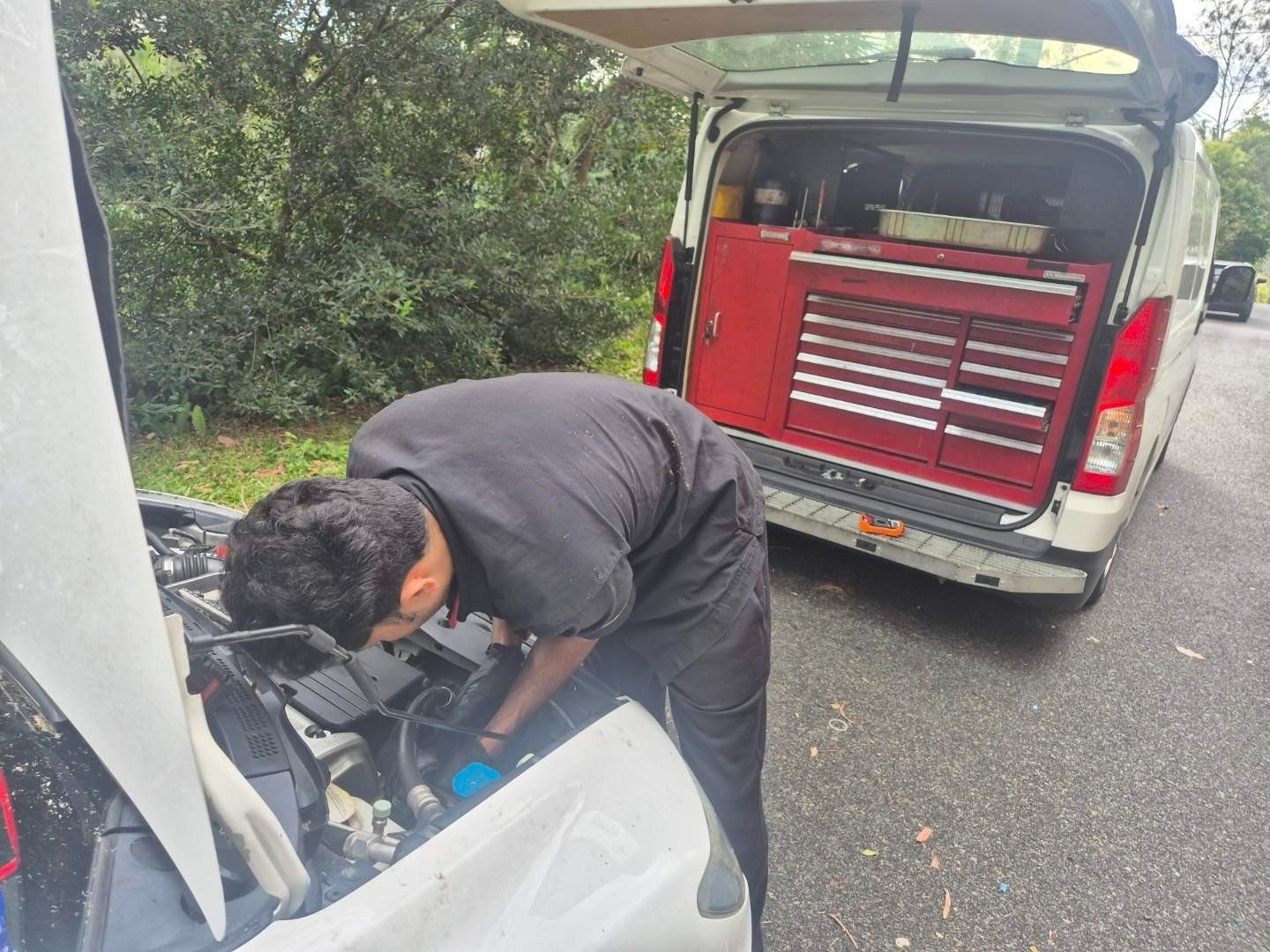 Mechanic working on car engine outdoors, van with tools open nearby.