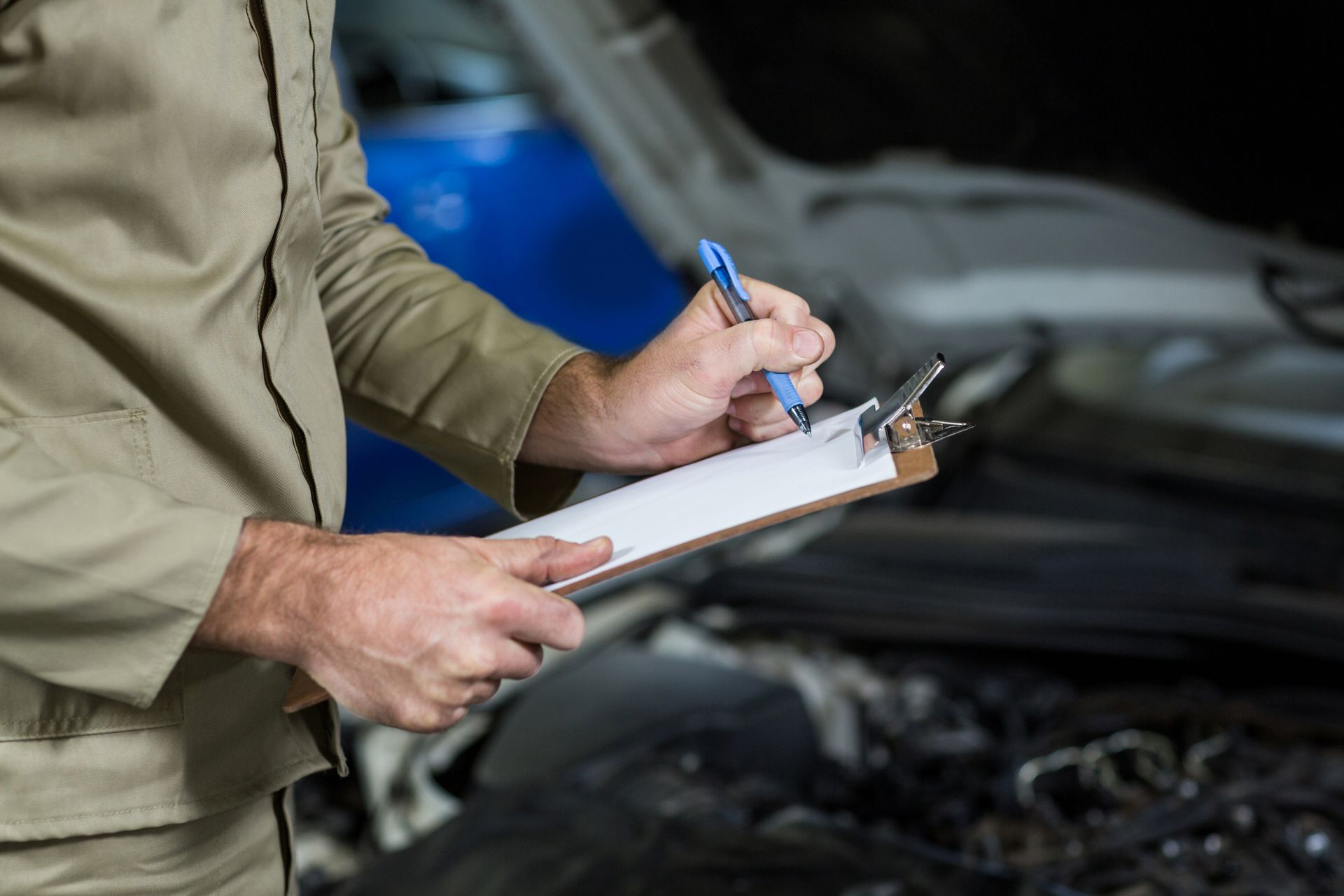 Mechanic in work uniform holding clipboard and pen, inspecting a car engine.