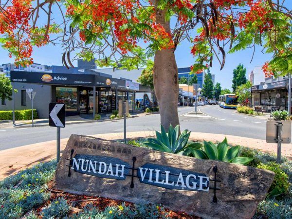 Nundah Village sign with a street view featuring shops, trees with red flowers, and a clear blue sky.