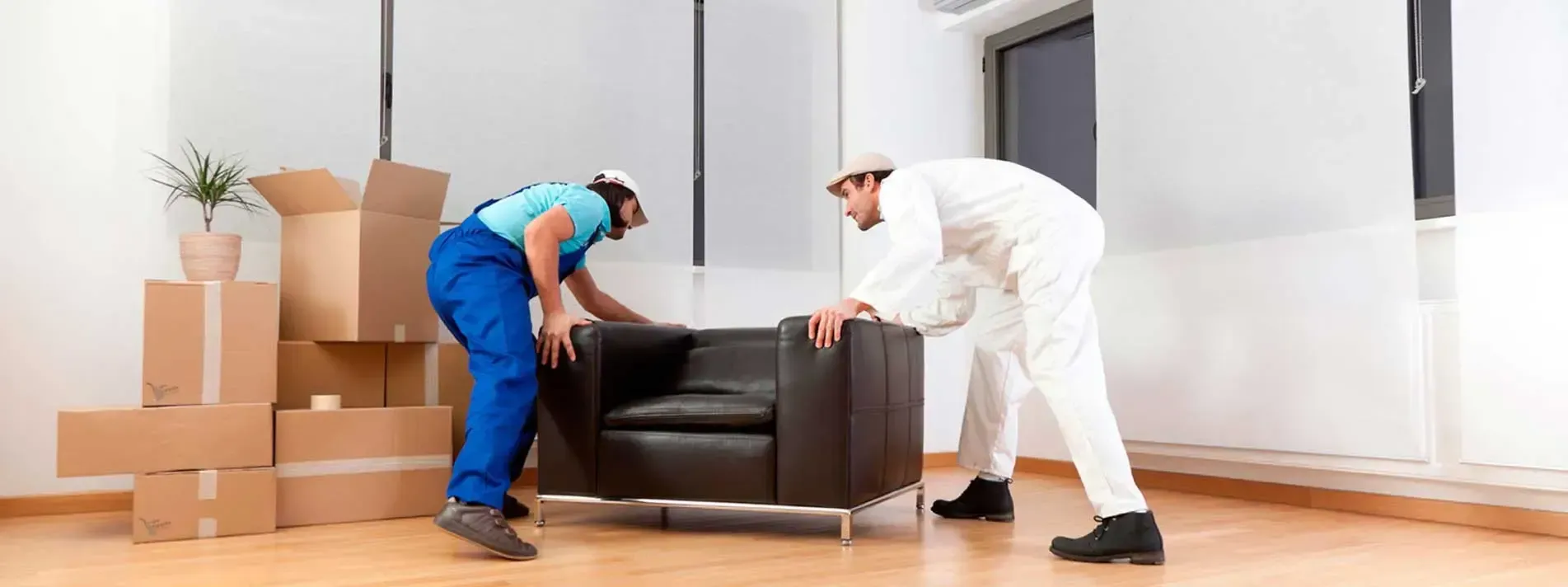 Dos operarios vestidos de trabajo levantan un sillón oscuro en una habitación con cajas de cartón.
