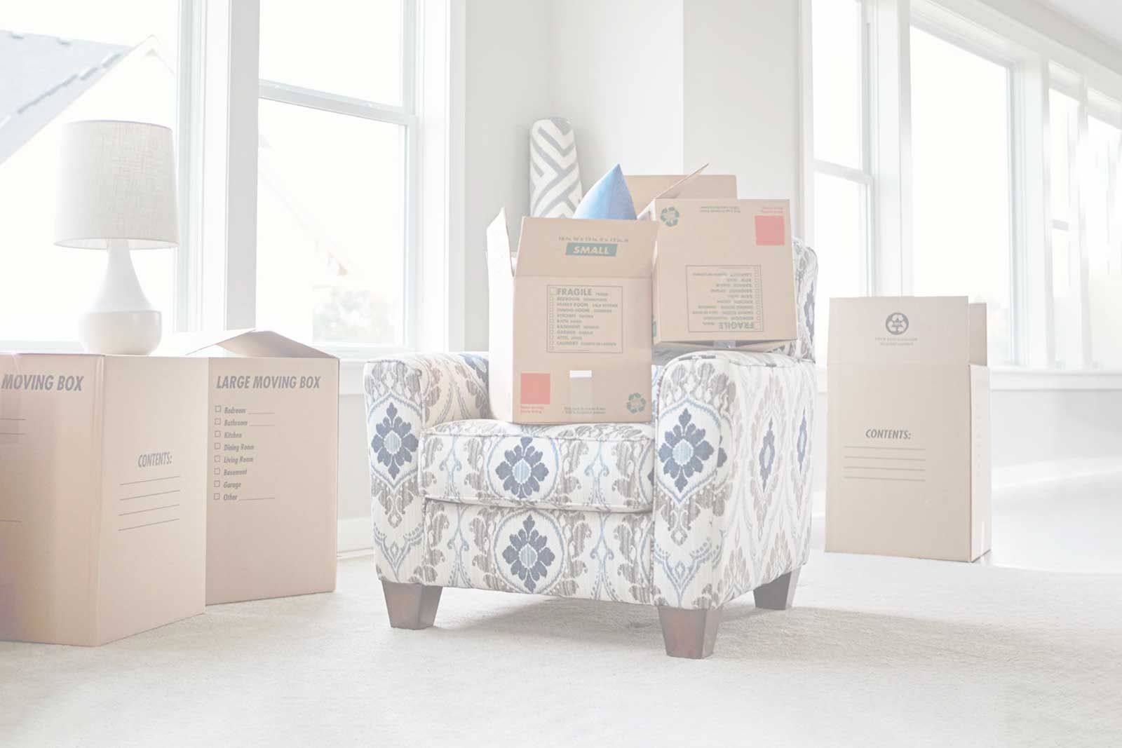 Cajas apiladas en un sillón estampado en una habitación luminosa, listas para ser trasladadas.