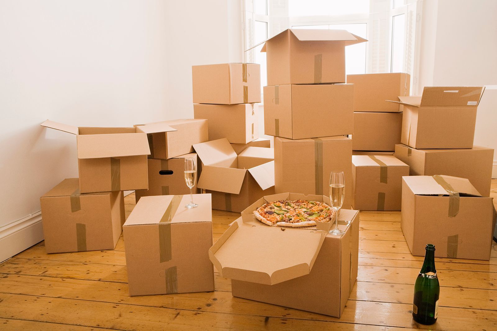 Cajas de cartón apiladas en una habitación con pizza, champán y un suelo de madera desnudo; día de mudanza.