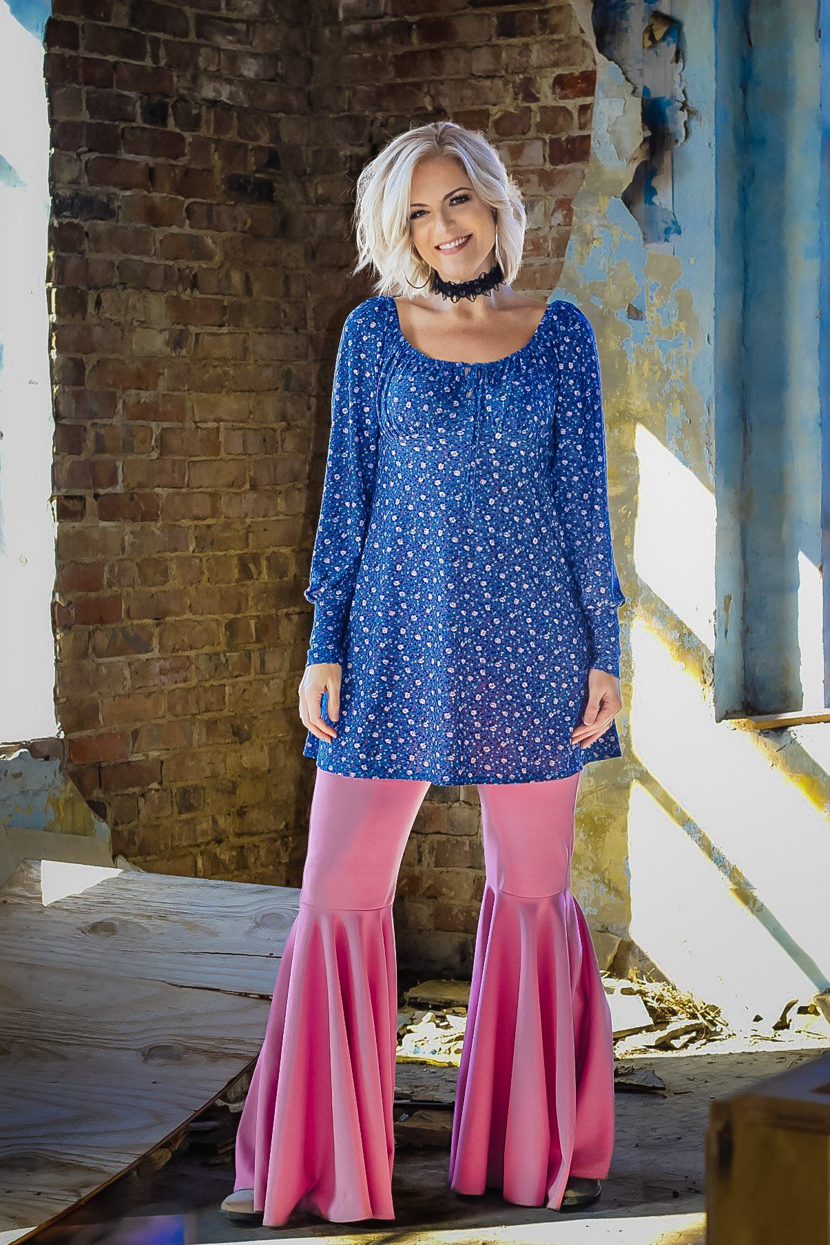 A woman in a blue top and pink pants is standing in front of a brick wall.