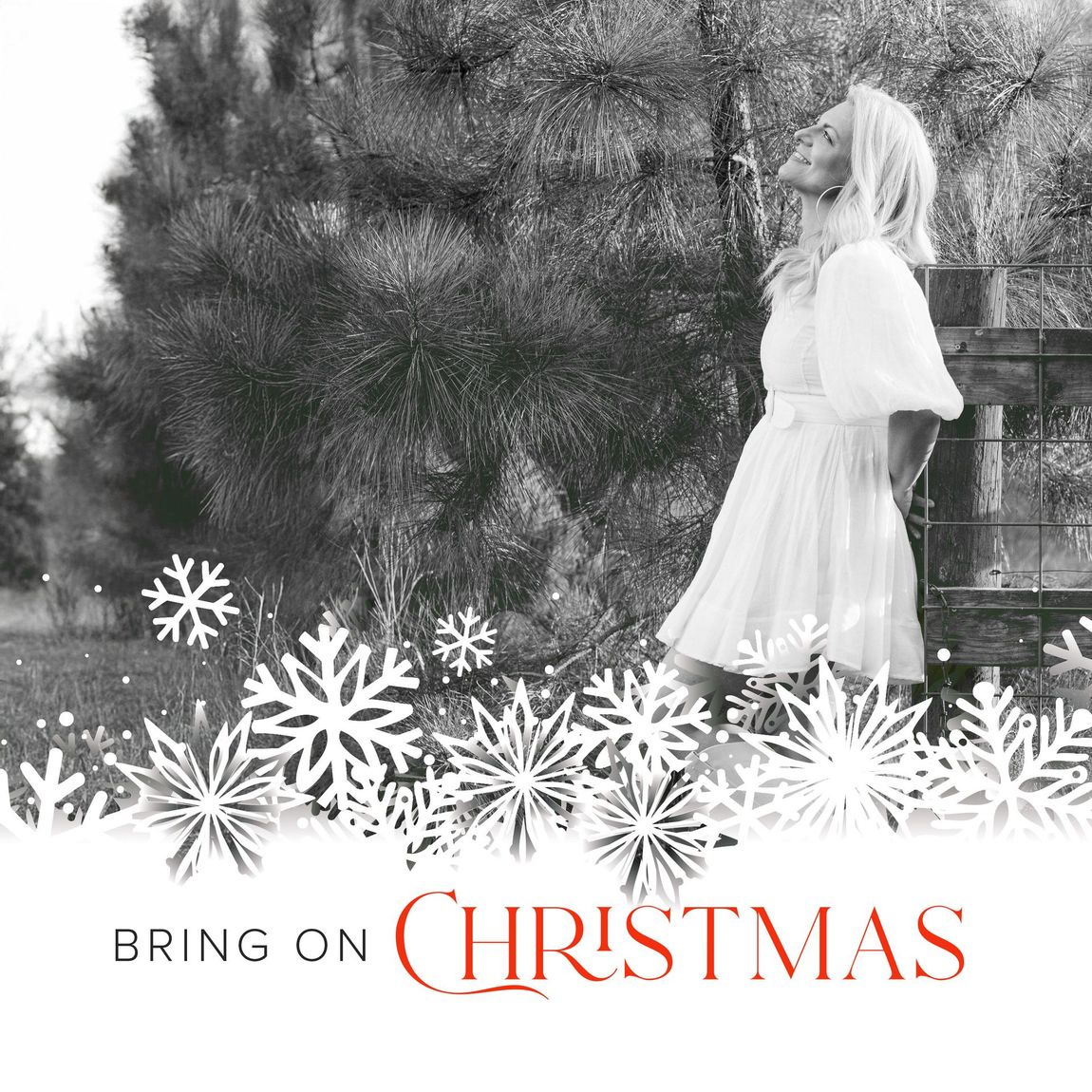 Bring On Christmas - EP Album cover by Delnora, leaning against a fence in a white dress with snowflakes and Christmas Trees in the background looking up to sky smiling