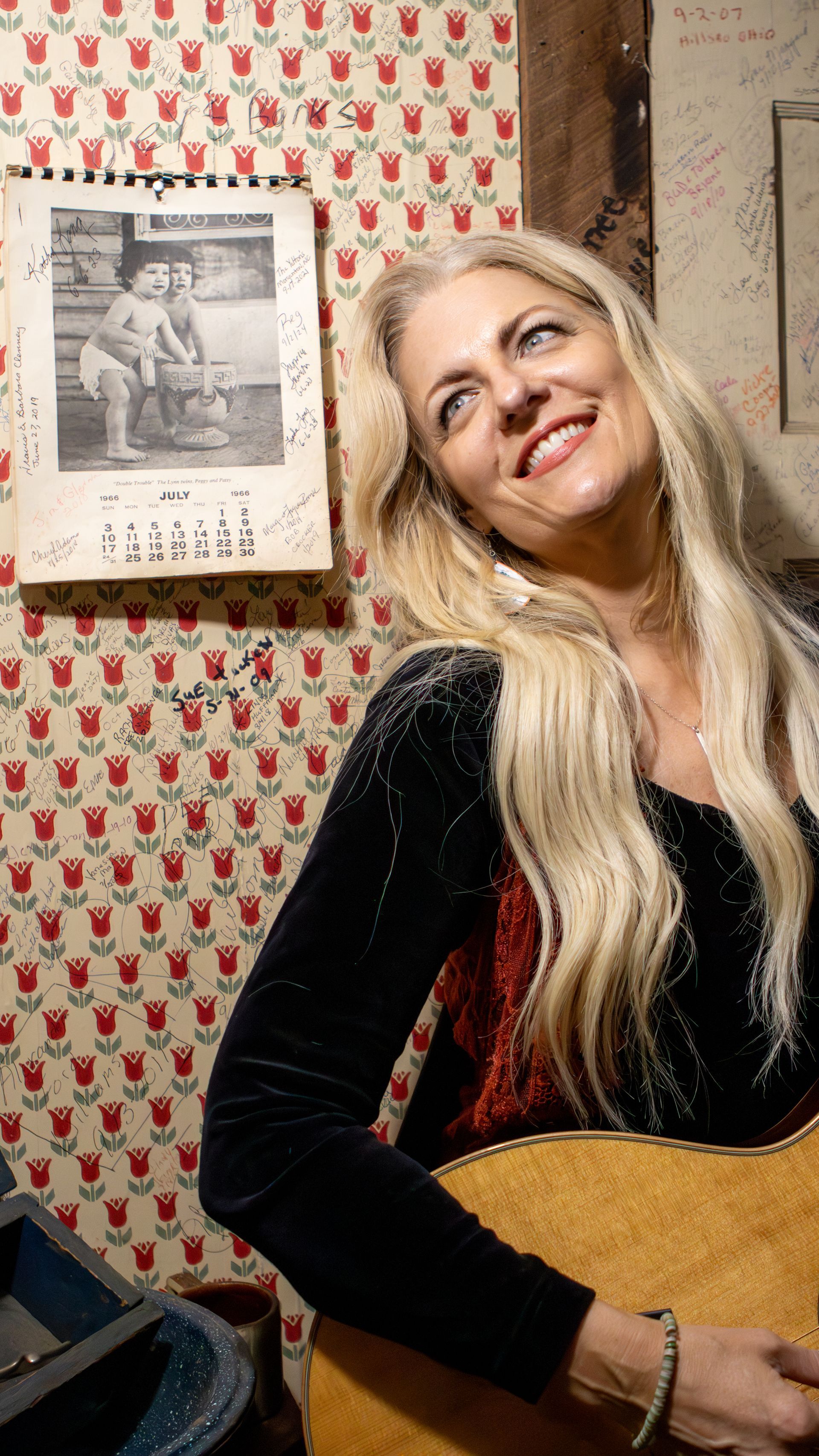 A woman is holding a guitar in front of a wall with a picture on it.