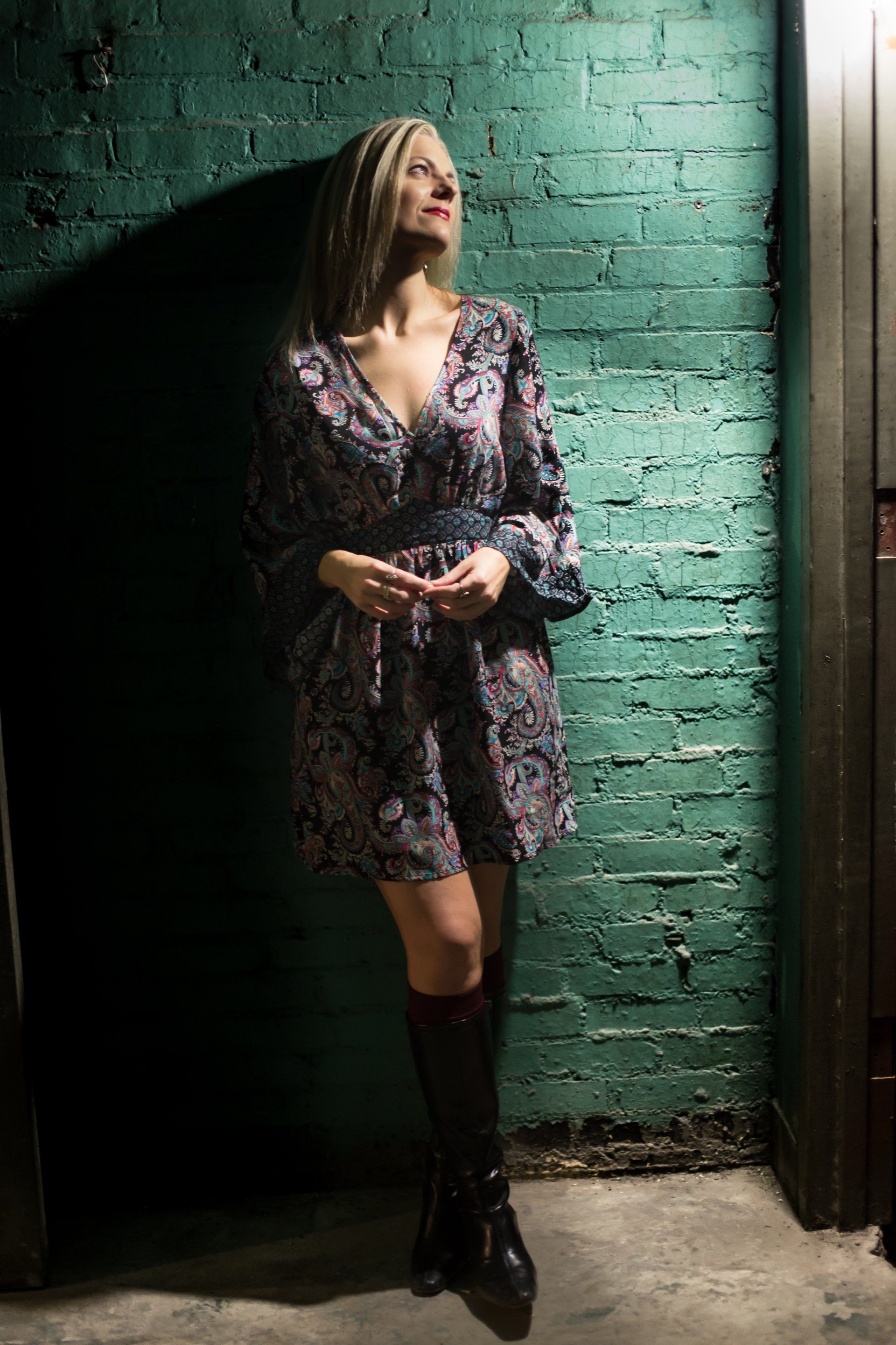 A woman in a dress and boots is standing in front of a green brick wall.