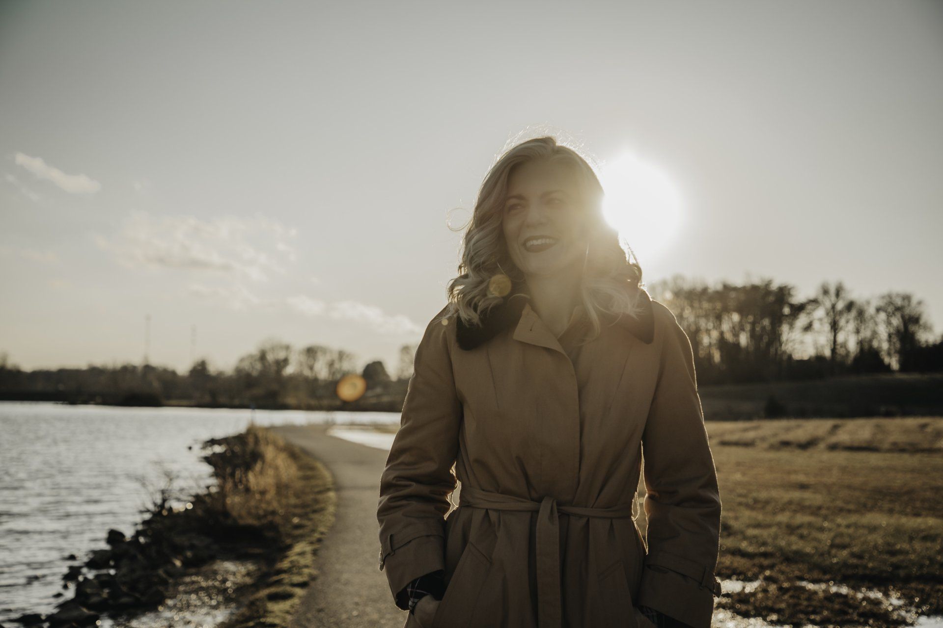 A woman in a trench coat is standing next to a body of water.