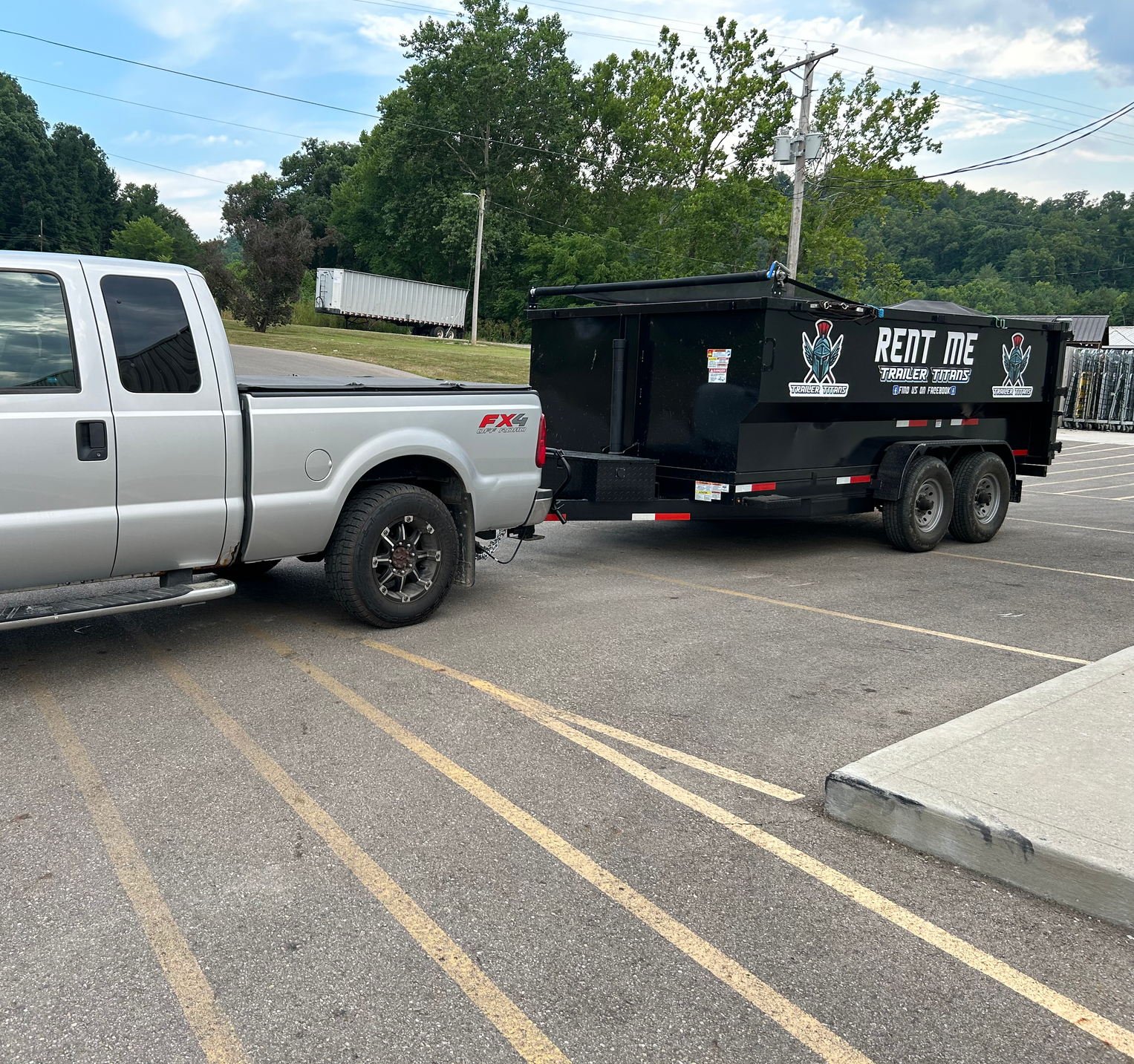 Silver pickup truck towing a black dumpster trailer in a parking lot.