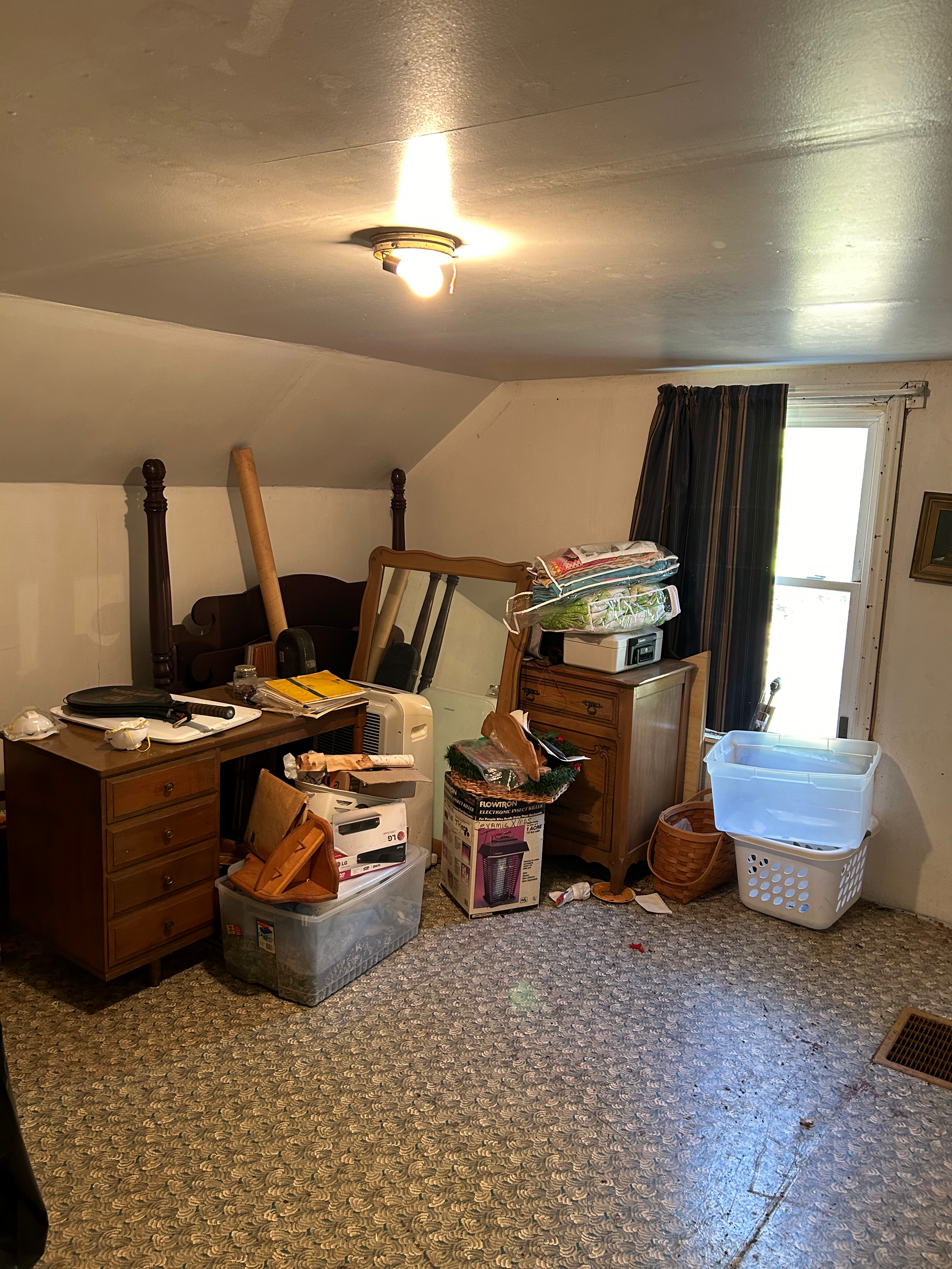 Messy attic room with furniture, boxes, and a window. Dark curtain, light fixture on ceiling, patterned floor.