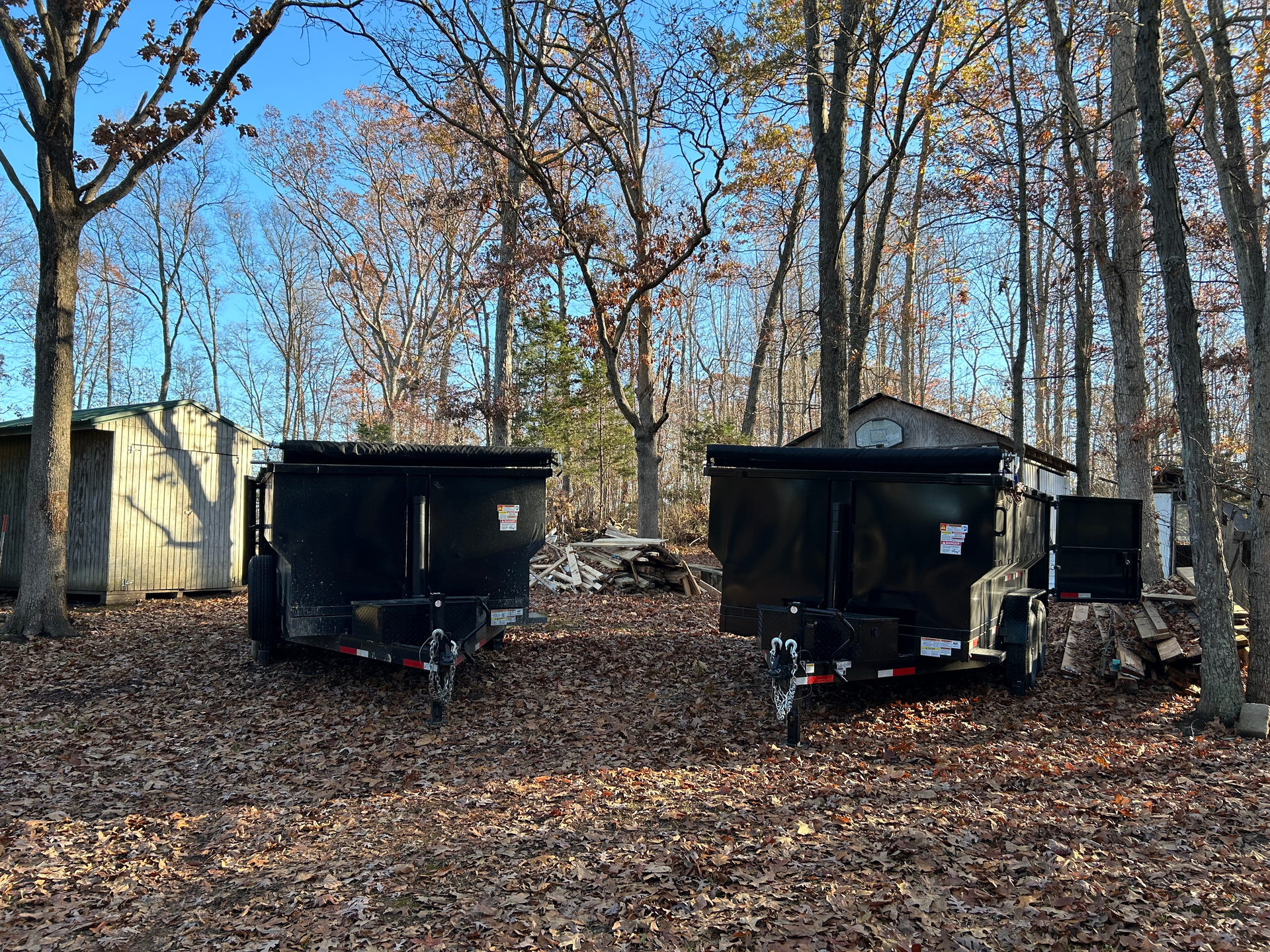 Two black dumpsters on trailers in a wooded area with fallen leaves and bare trees.