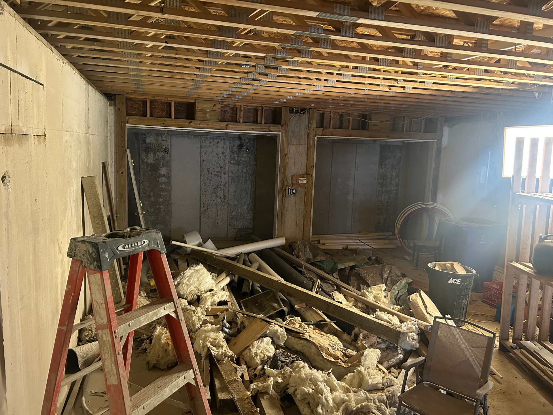 Interior demolition with ladder, debris, exposed ceiling joists and two wall openings.