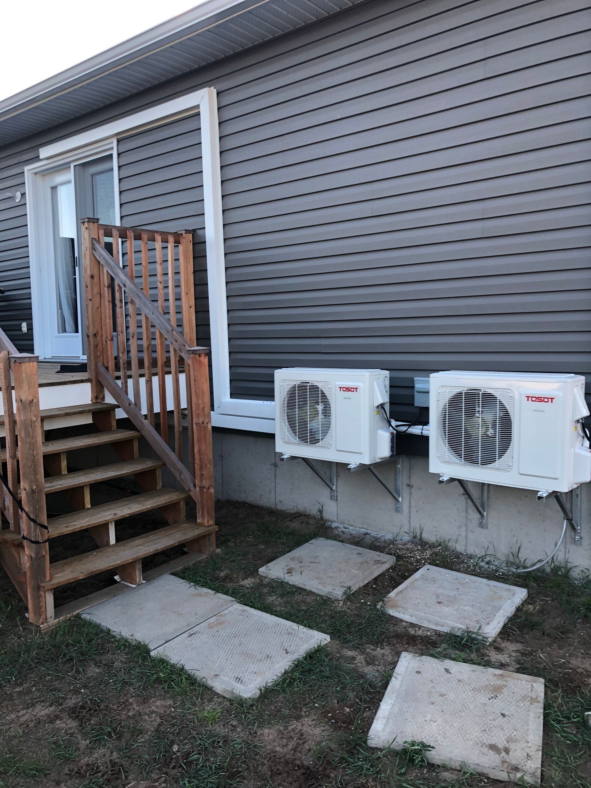 Deux unités de climatisation extérieures blanches, fixées sur un mur de maison recouvert de vinyle gris, à côté d'un escalier en bois.