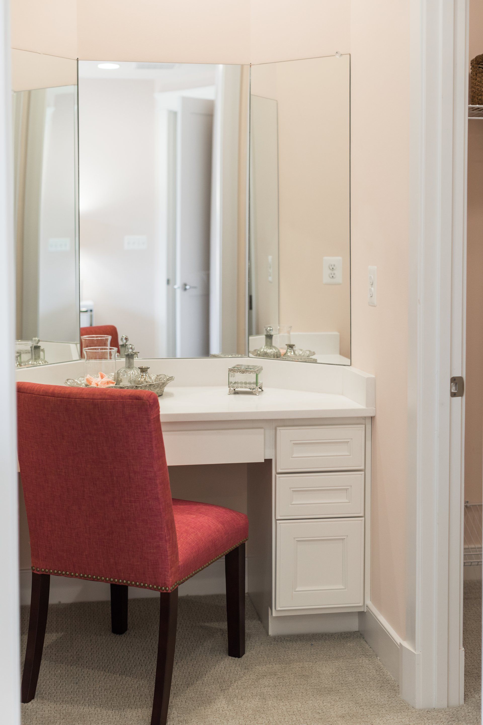 Vanity with red chair, triple mirror, and white drawers in a pink room.
